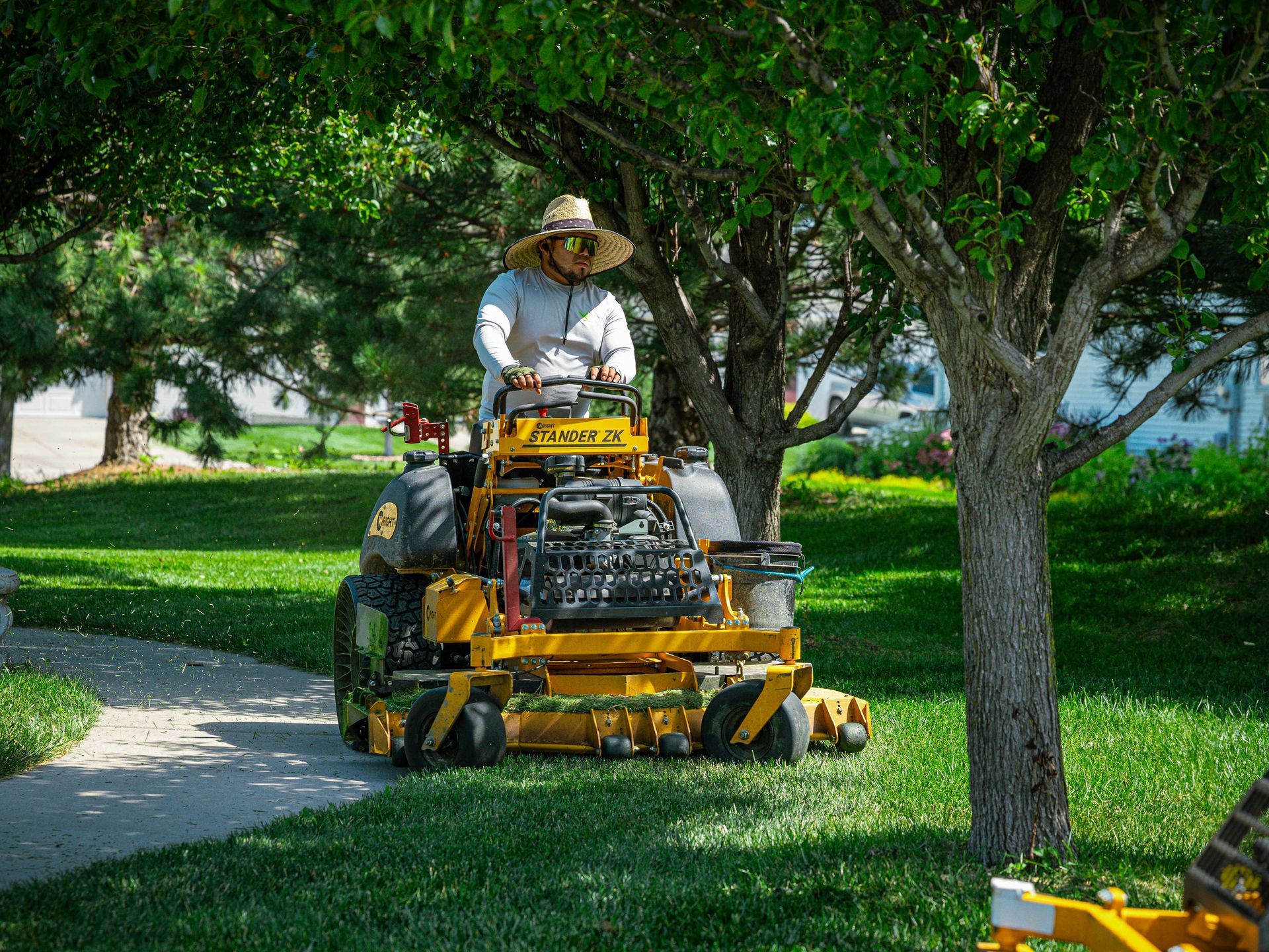 Worker mowing grass on a yellow riding mower in a tree-lined park beside a sidewalk