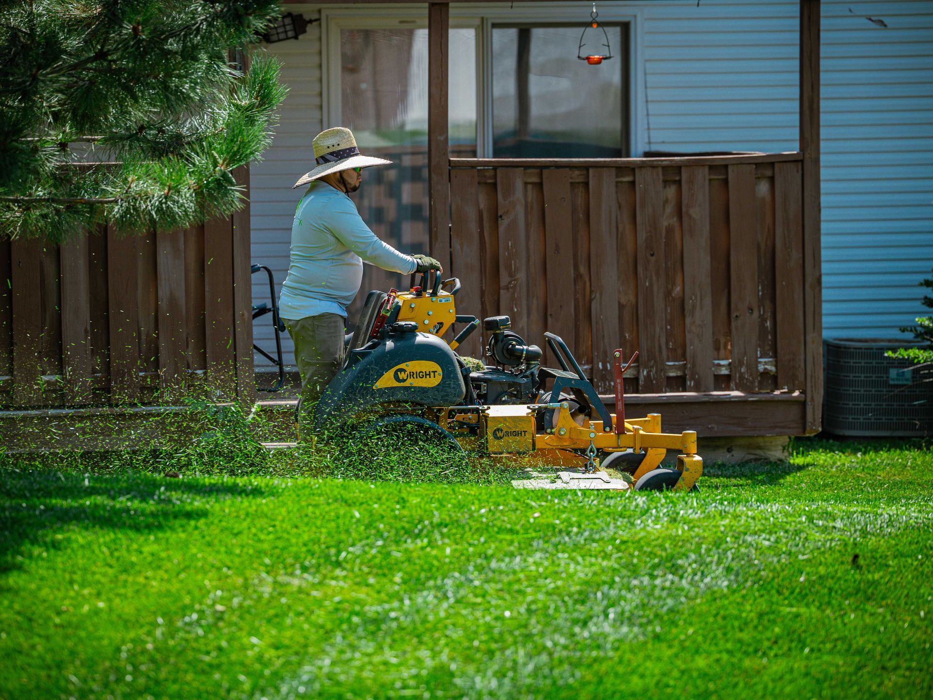 Person mowing a green lawn with a yellow riding mower in a backyard beside a wooden fence.