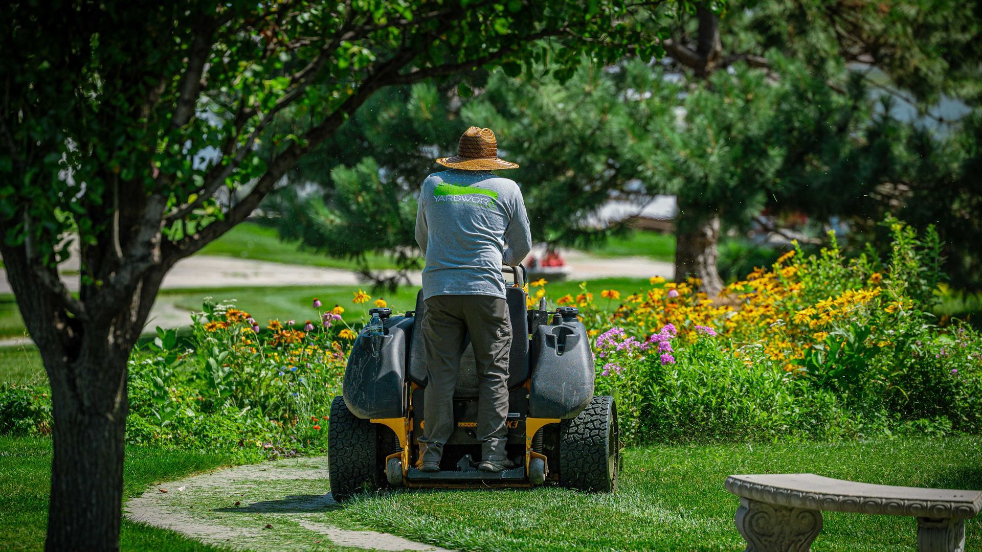 Person mowing a grassy park path with a riding lawn mower, surrounded by green trees and yellow flowers
