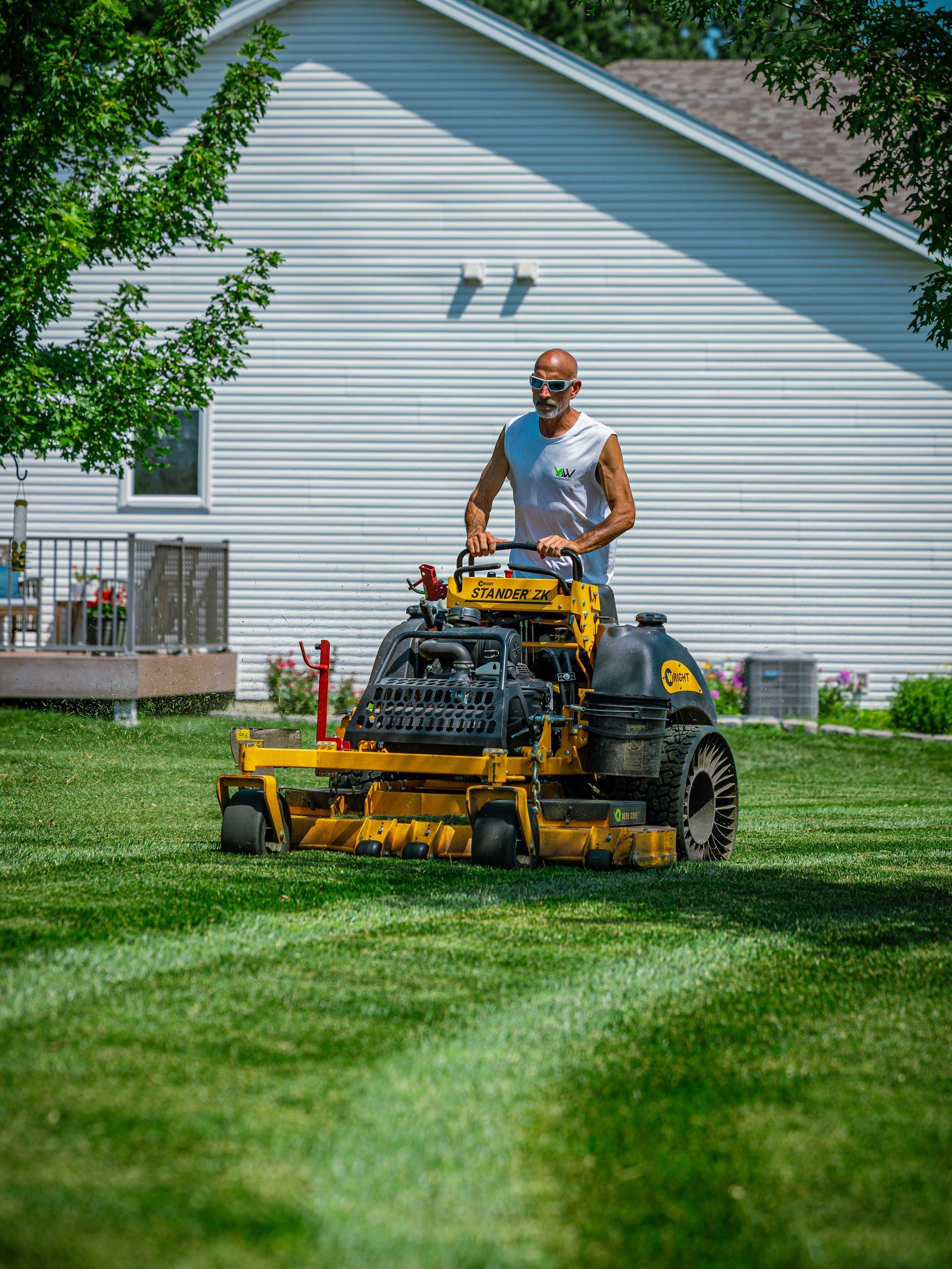 Man mowing a green lawn with a yellow riding mower in front of a white house
