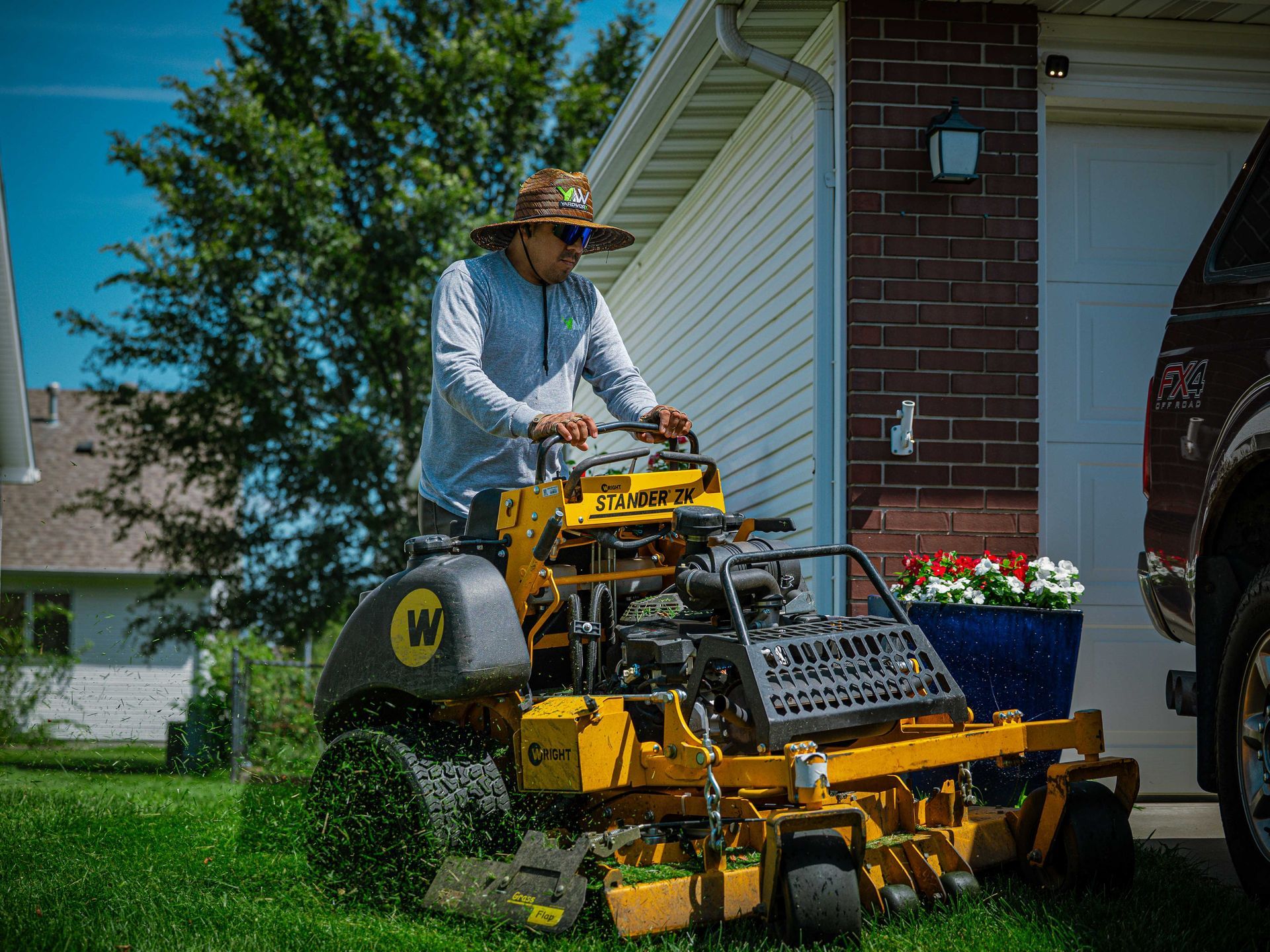 Worker operating a yellow lawn mower beside a house with trimmed green grass and flowers