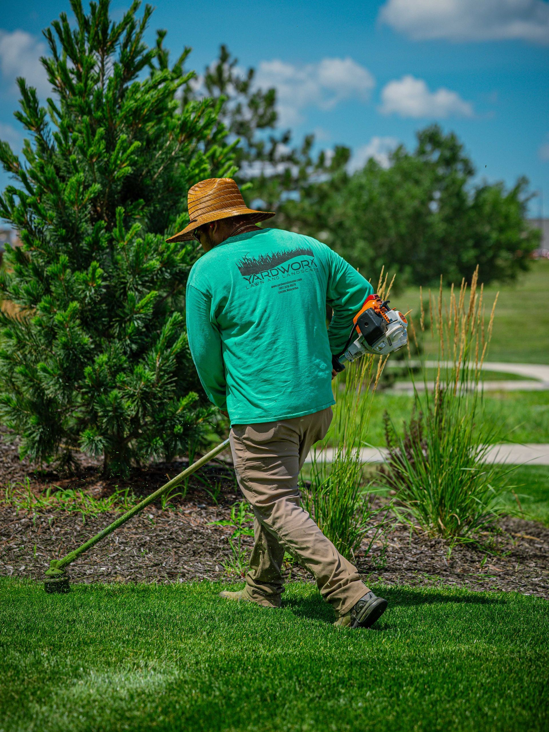 Person in a straw hat using a string trimmer on a grassy lawn near shrubs and trees