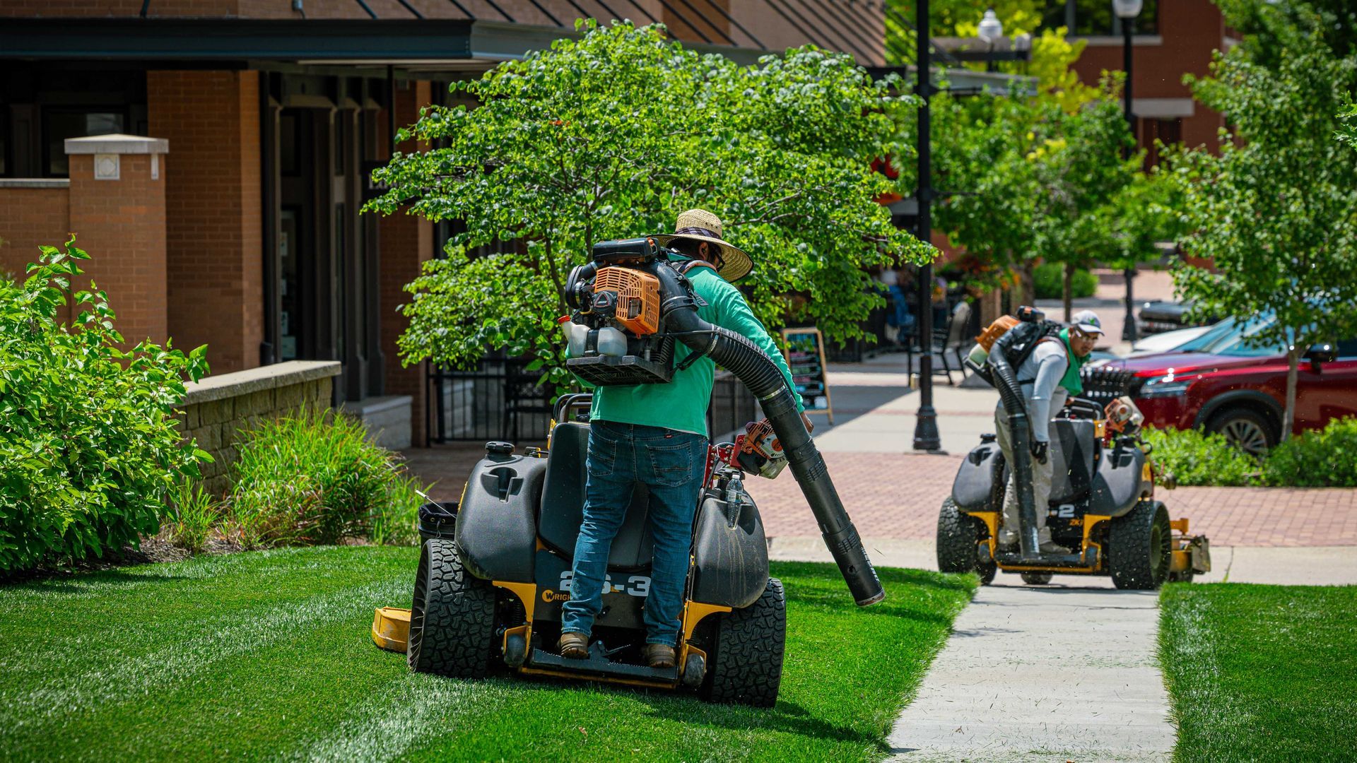 Two landscapers mowing and trimming a green lawn beside a sidewalk outside brick buildings