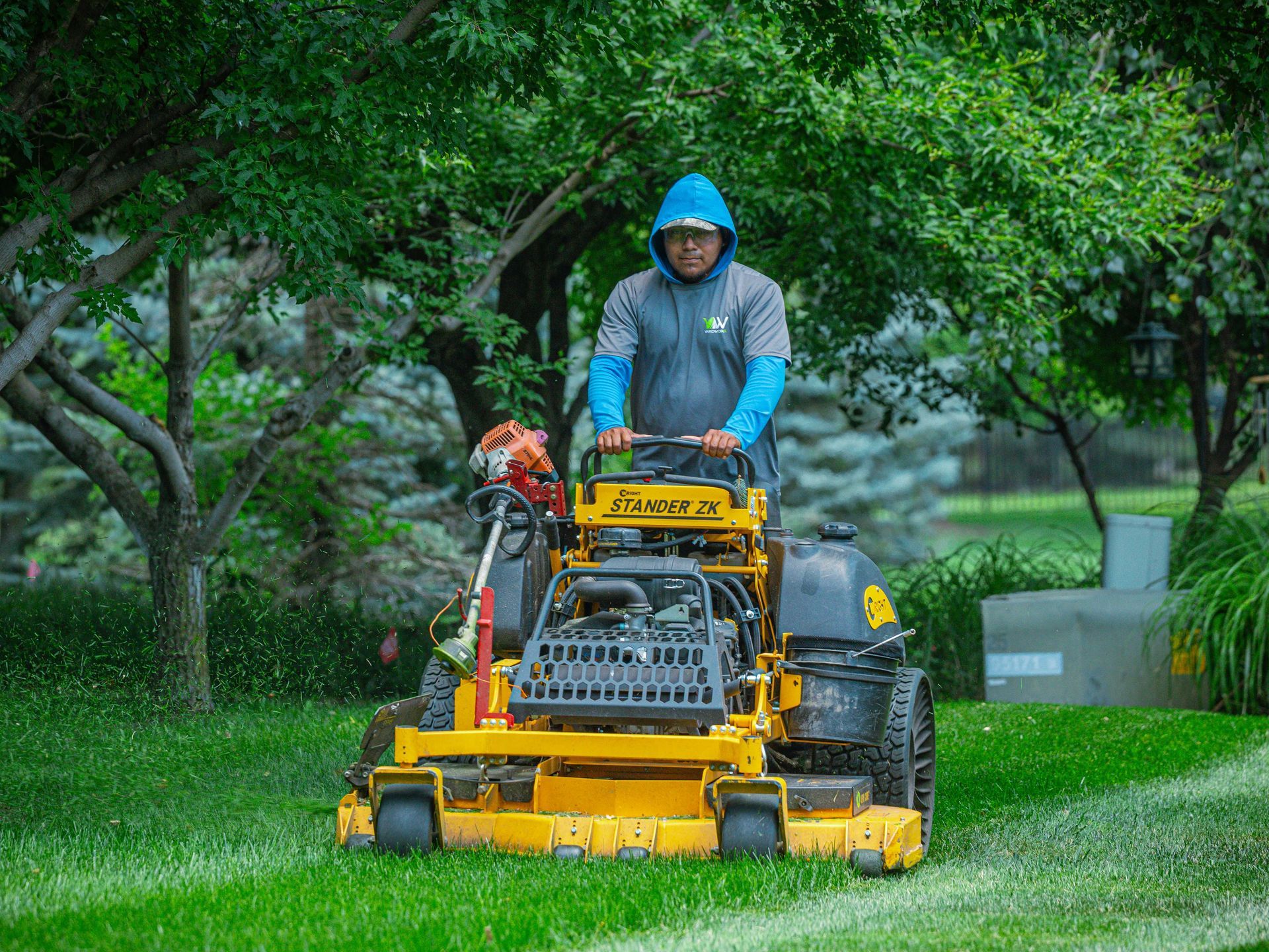 Person operating a yellow lawn mower in a lush green garden, wearing a blue hooded rain jacket.