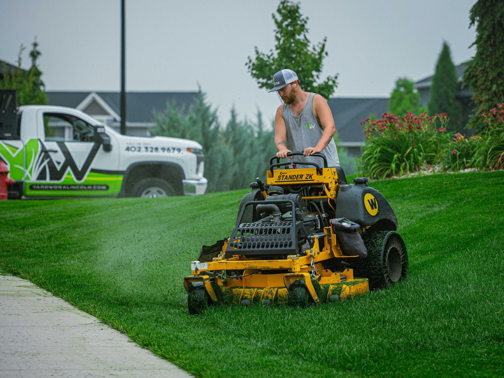 Worker mowing a wet lawn with a yellow riding mower near a driveway on a rainy day