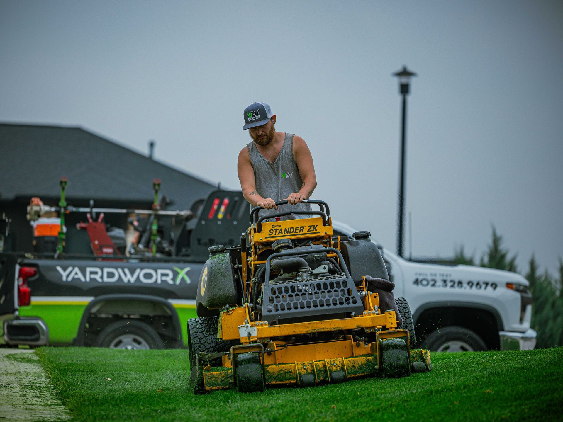 Person mowing grass on a yellow ride-on mower at an outdoor event with trucks in the background