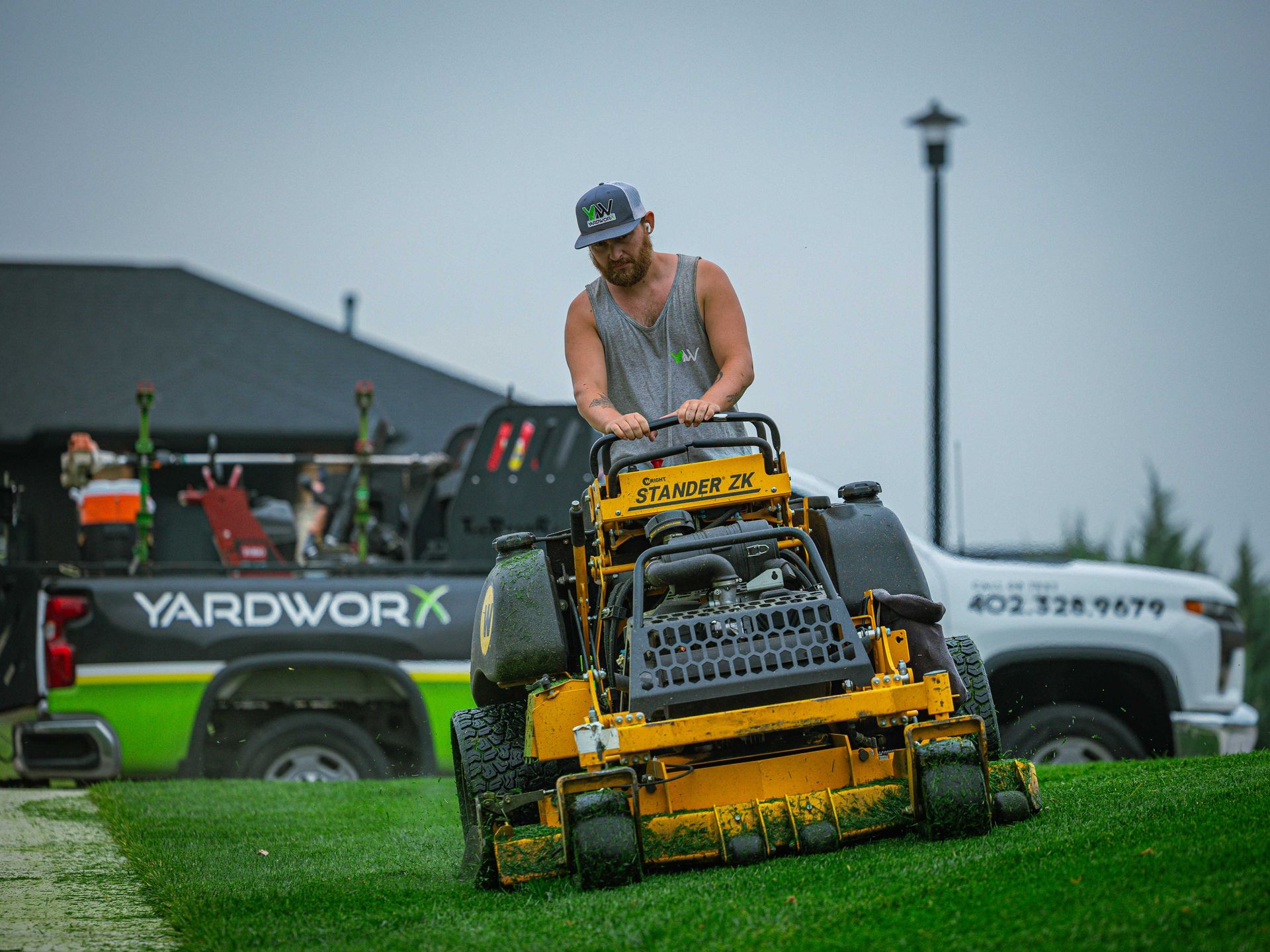 Man mowing a wet lawn on a yellow riding mower with yardwork vehicles in the background