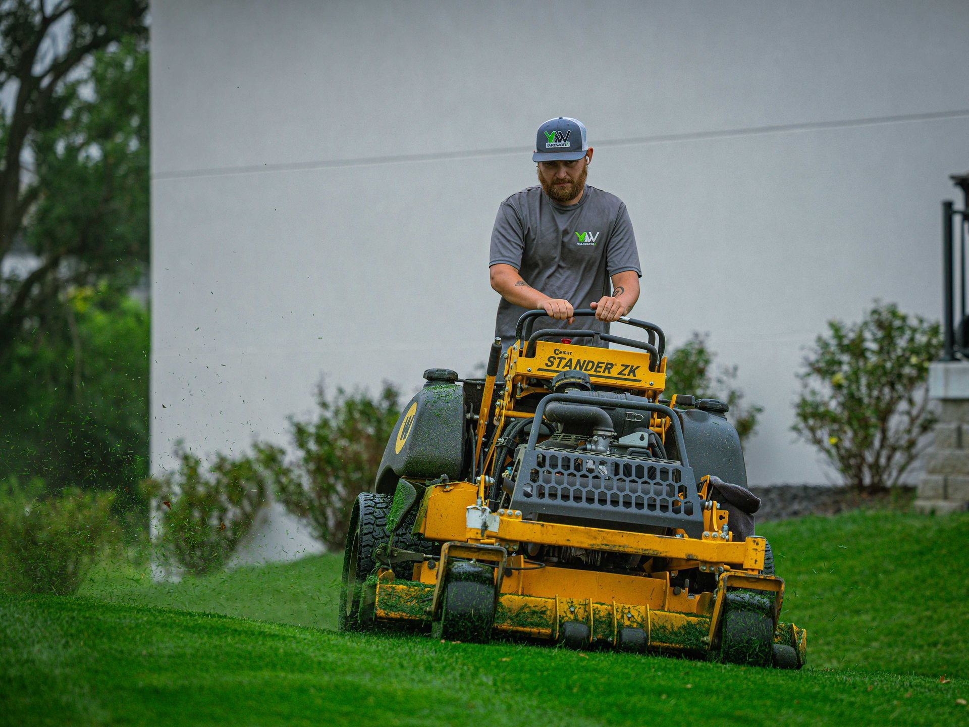 Man mowing a bright green lawn with a yellow riding mower near a gray building