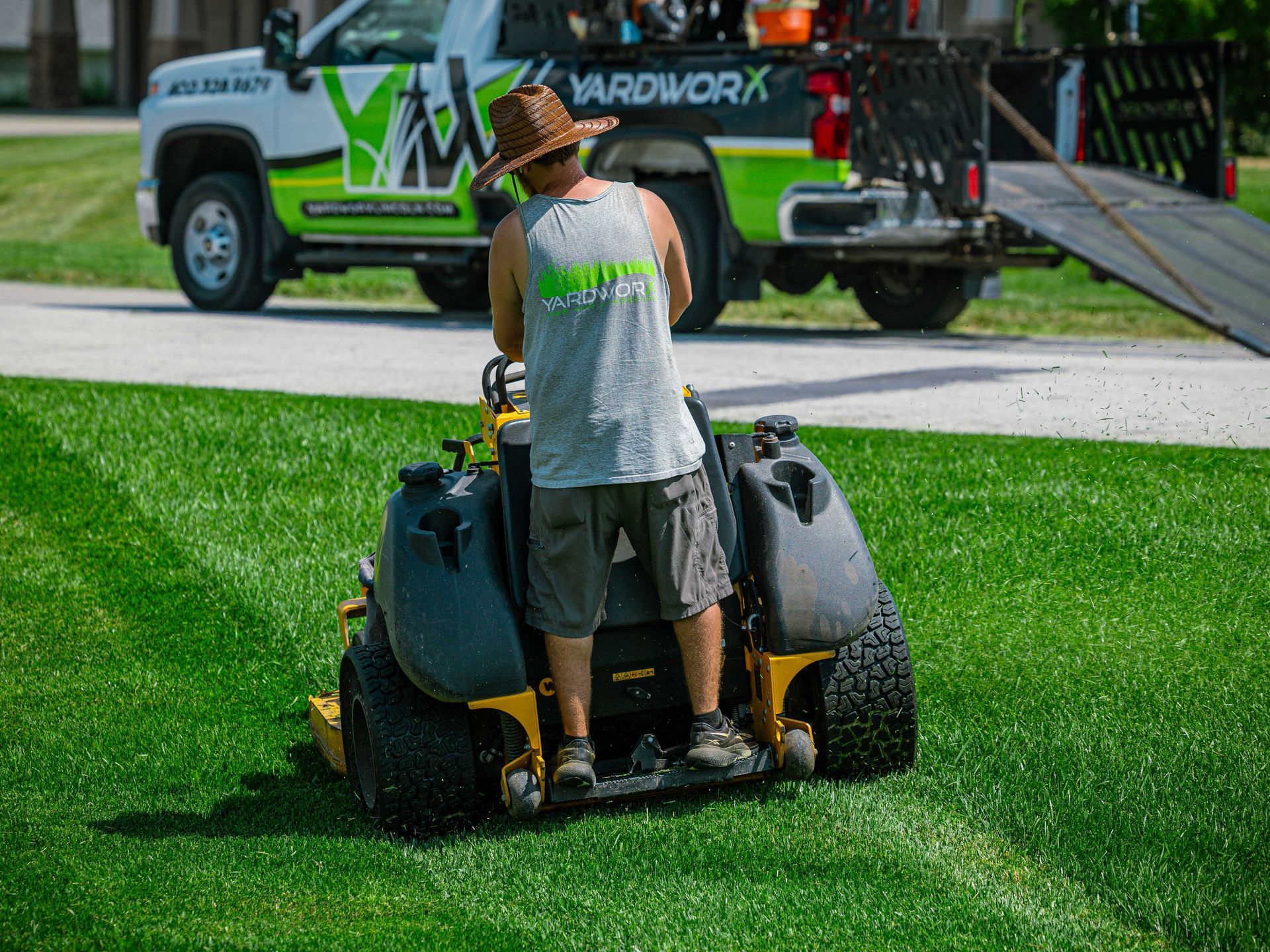 Worker operating a green zero-turn mower on a grassy lawn near a service truck.
