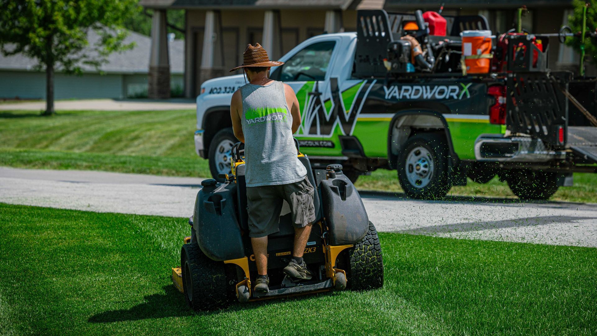 Worker mowing a green lawn beside a black-and-green service truck parked on a street