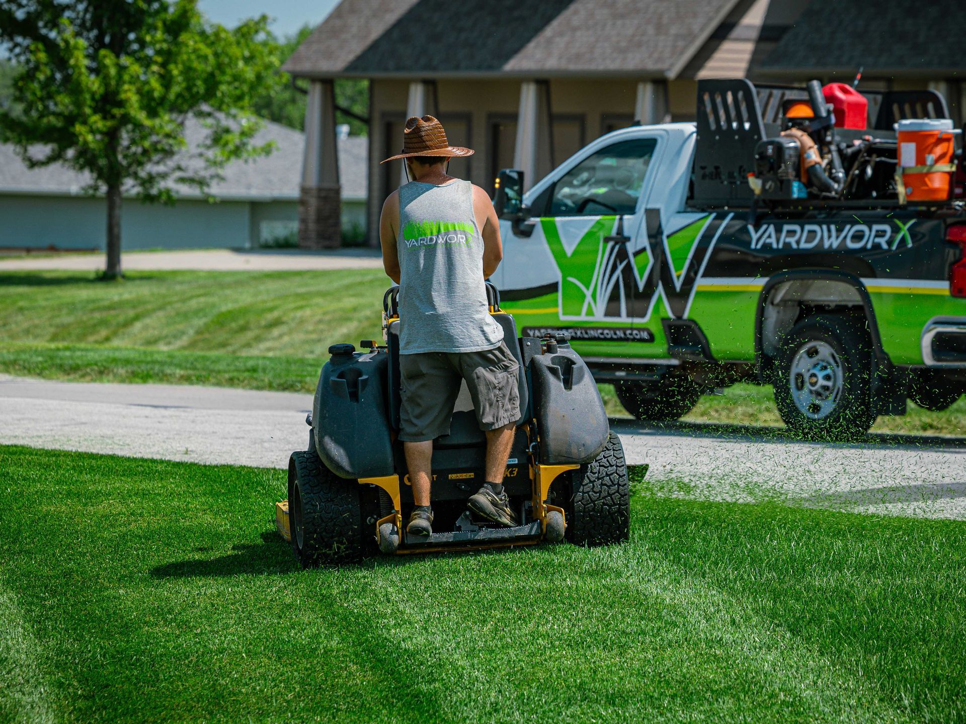 Person mowing a green lawn with a push mower beside a landscaping truck in a suburban yard.