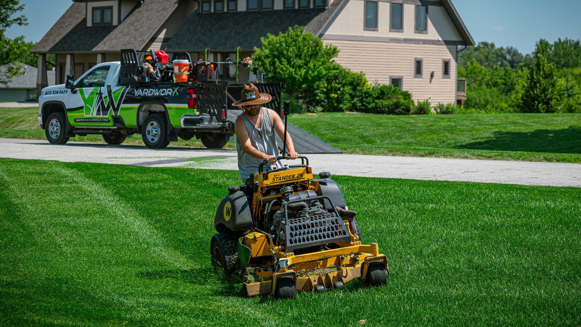Person mowing a lush green lawn with a yellow riding mower in front of a suburban house