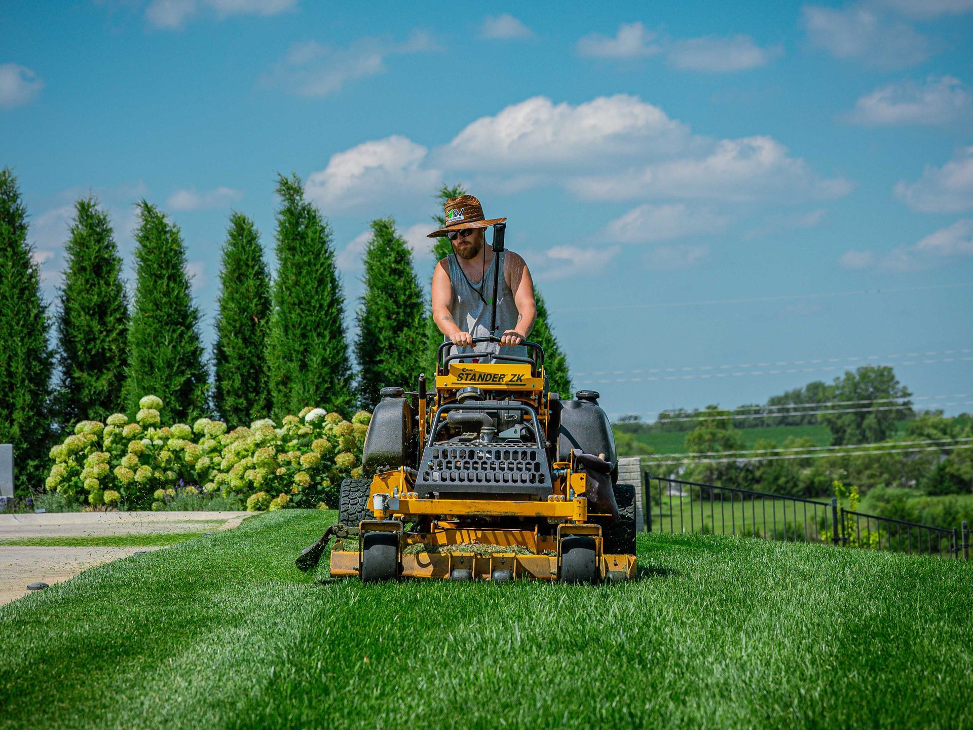 Person mowing a lush green lawn with a riding mower beside tall trees under a blue sky