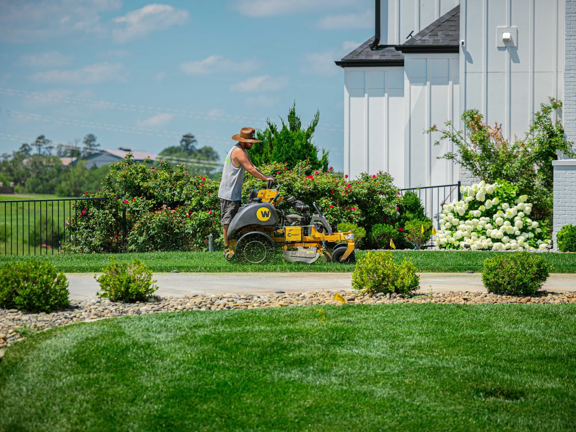 Person mowing a lawn beside a white house and garden on a sunny day