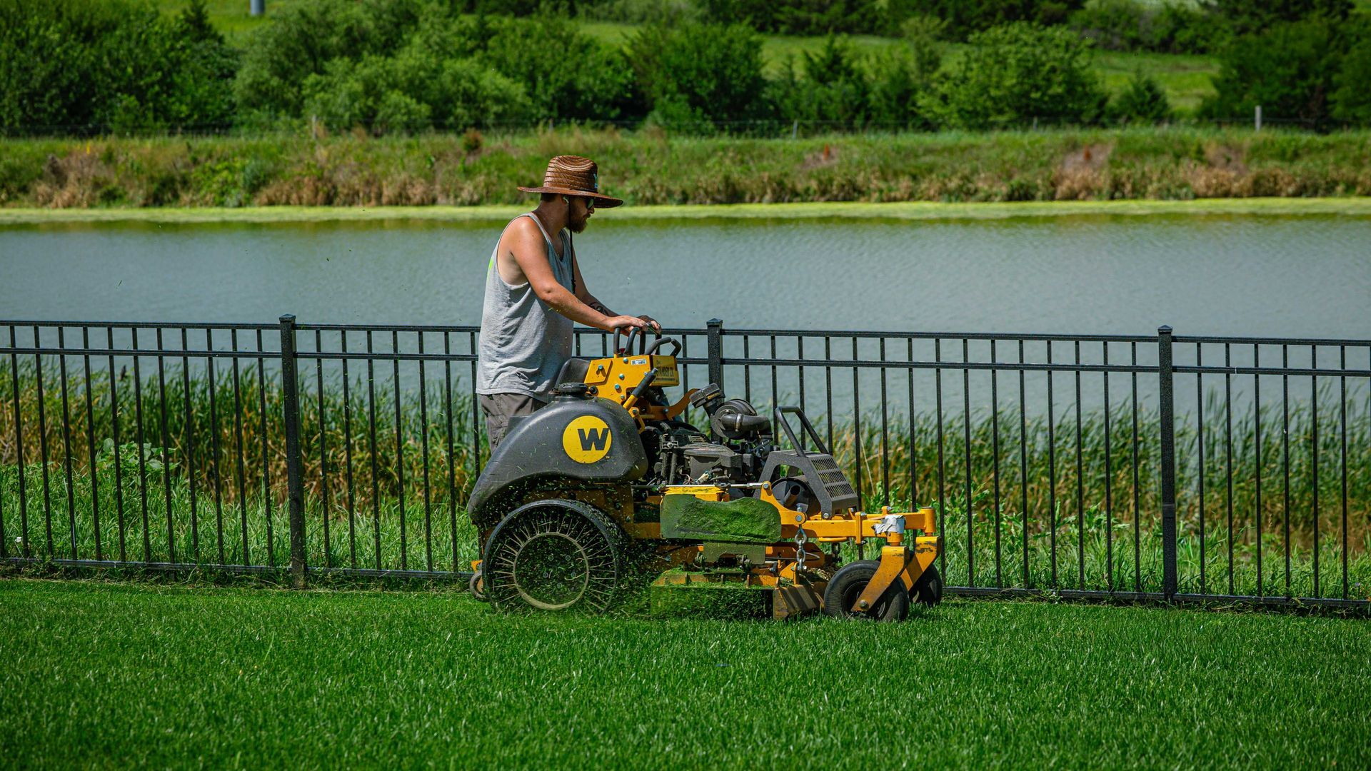 Person mowing bright green grass beside a lake with a yellow riding mower and black fence in the background