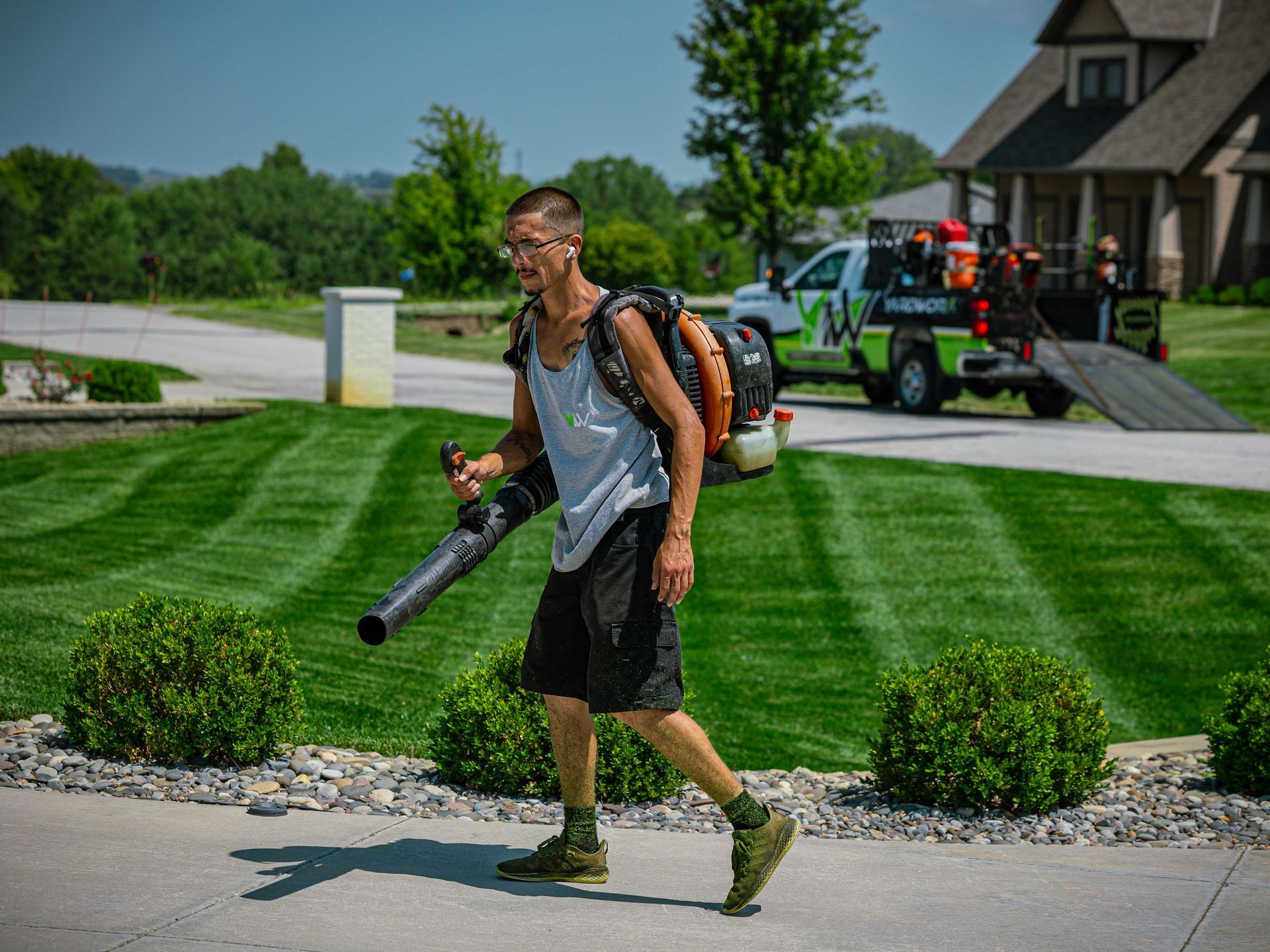 Person walking on a suburban sidewalk with a large leaf blower and backpack sprayer, lawns and house behind.