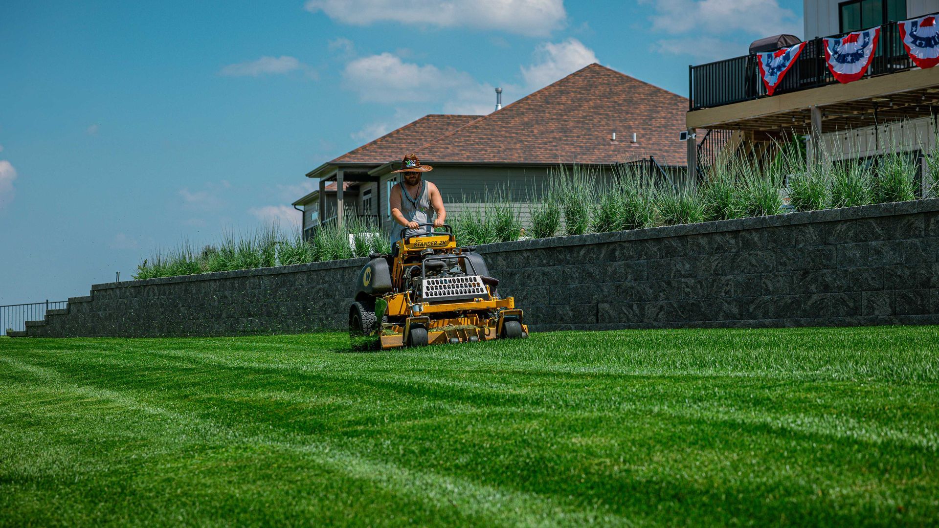 Person mowing a green lawn beside a house with a stone wall and tiled roof.