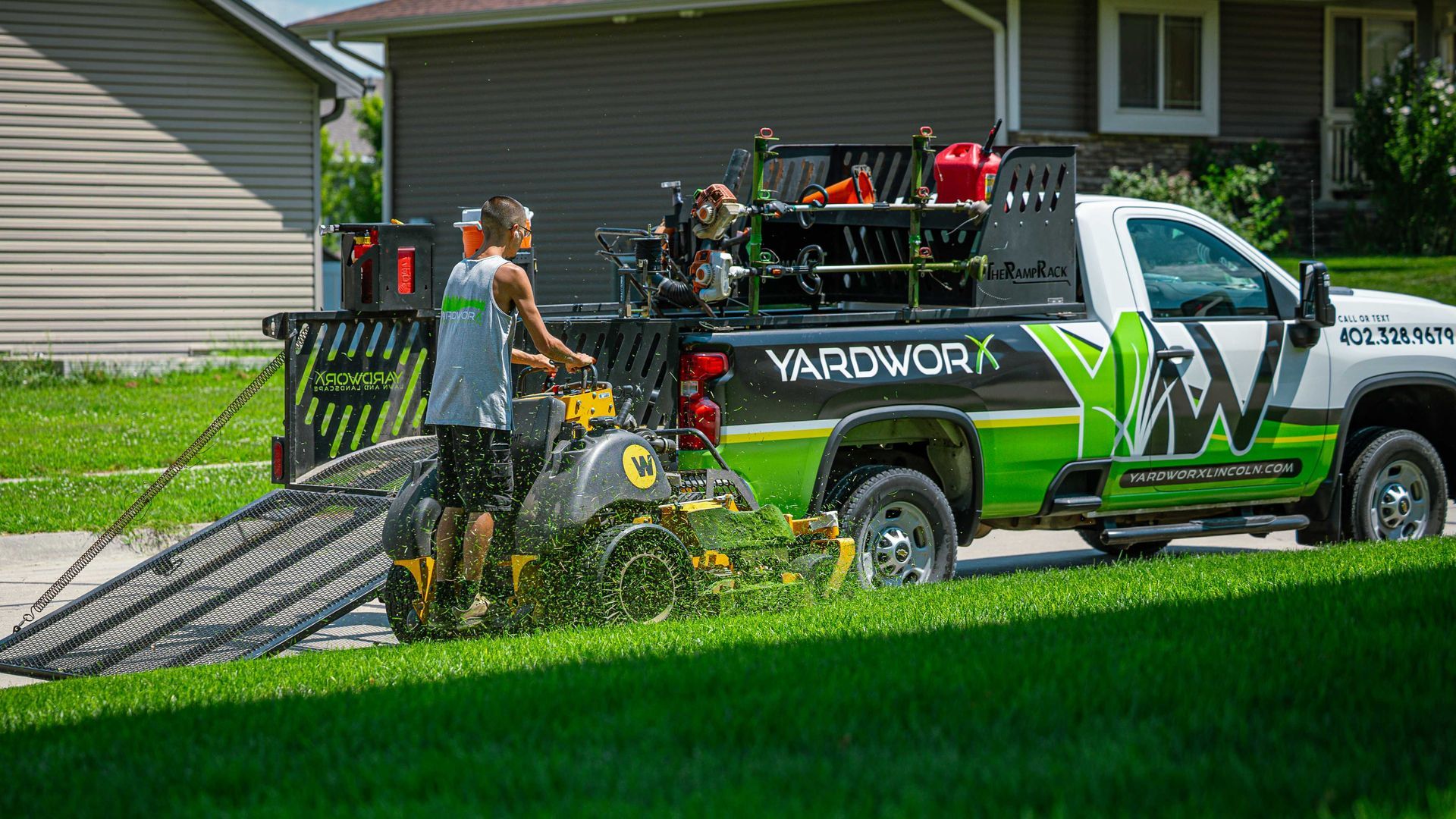 Landscaping crew loading a mower ramp onto a green and white YARDWORKS truck in a suburban yard
