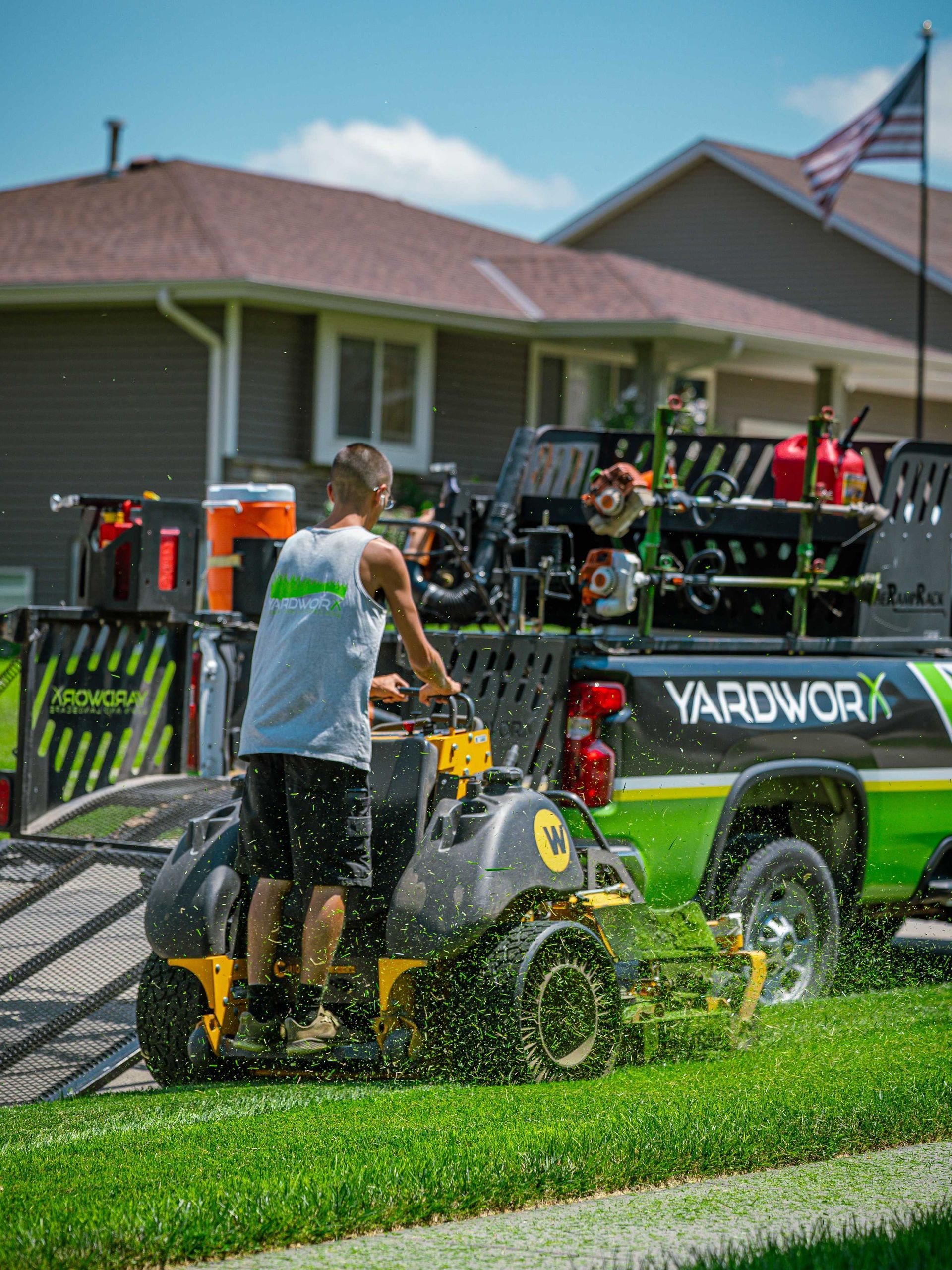 Person mowing a lawn in front of a house, pulling a black-and-yellow riding mower across wet grass.