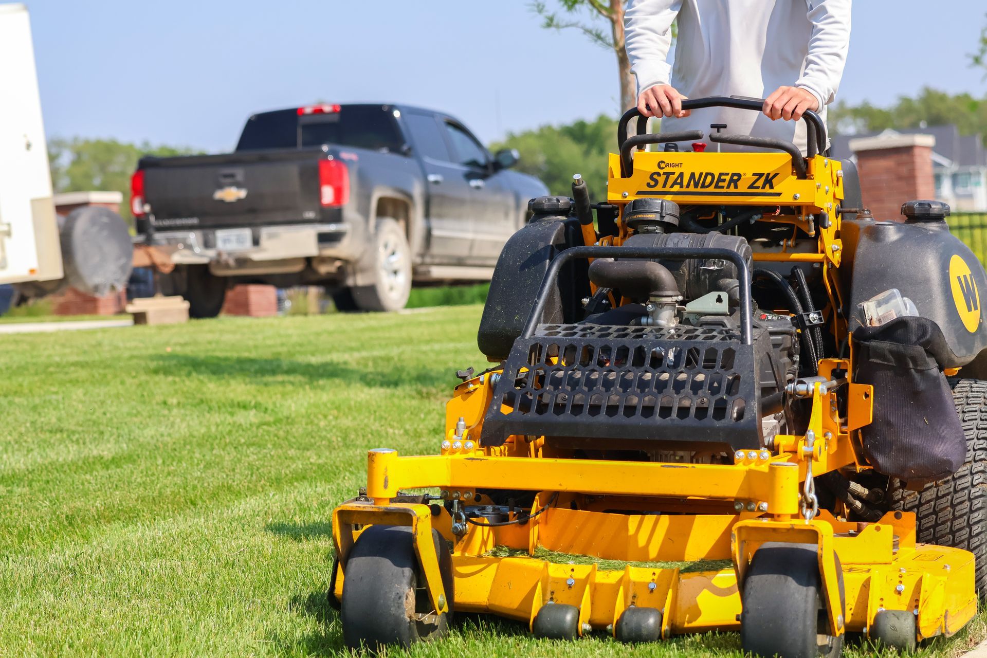 Yellow zero-turn mower on green grass, operator standing, truck in background.