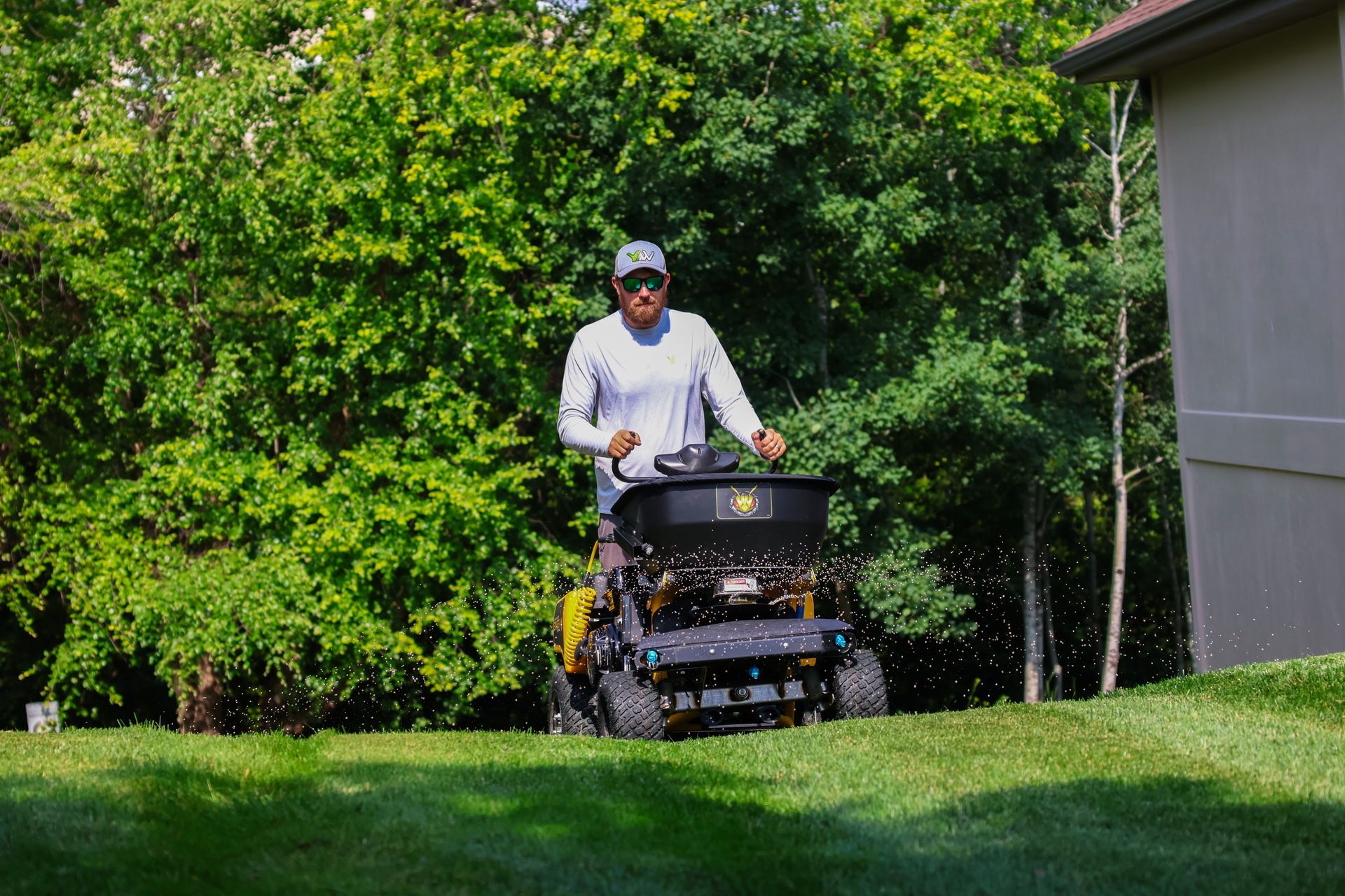Man operating a lawn spreader on a grassy lawn near trees and a building.