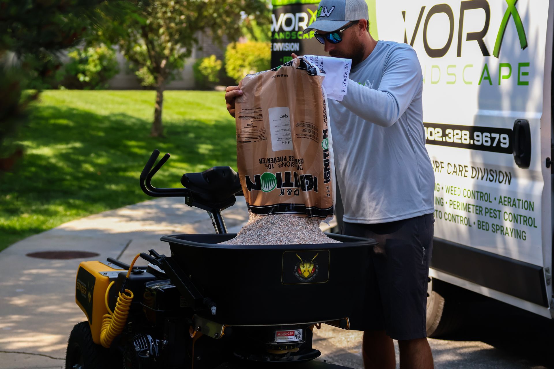 Man pouring fertilizer from bag into a spreader next to a vehicle with landscaping logo.