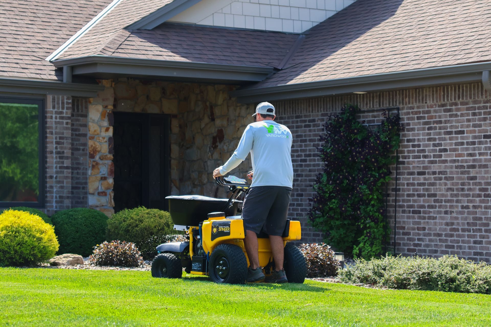 Man operating a yellow lawn spreader on a green lawn in front of a brick house.