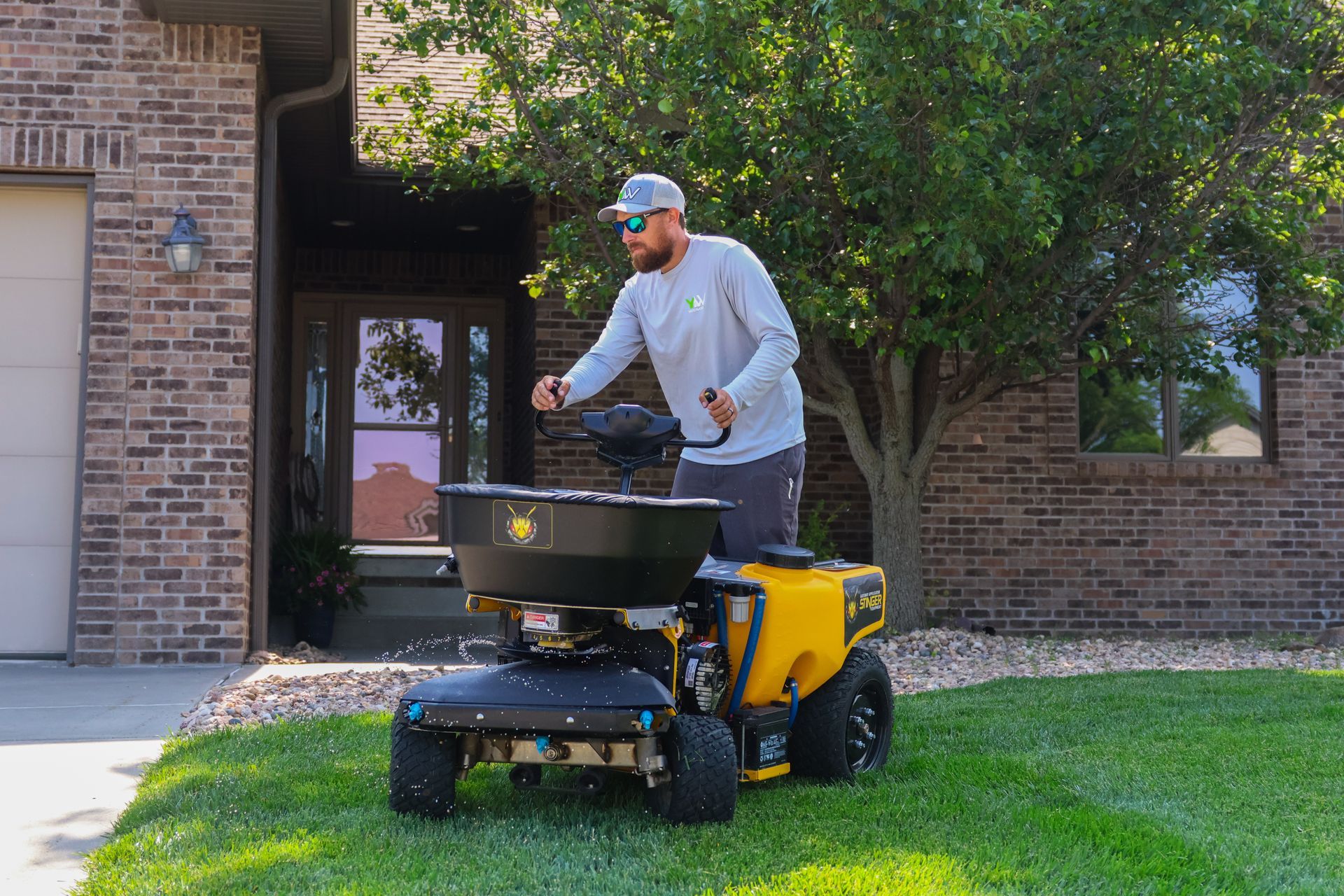 Man spreading fertilizer on lawn with a yellow machine in front of a brick house.