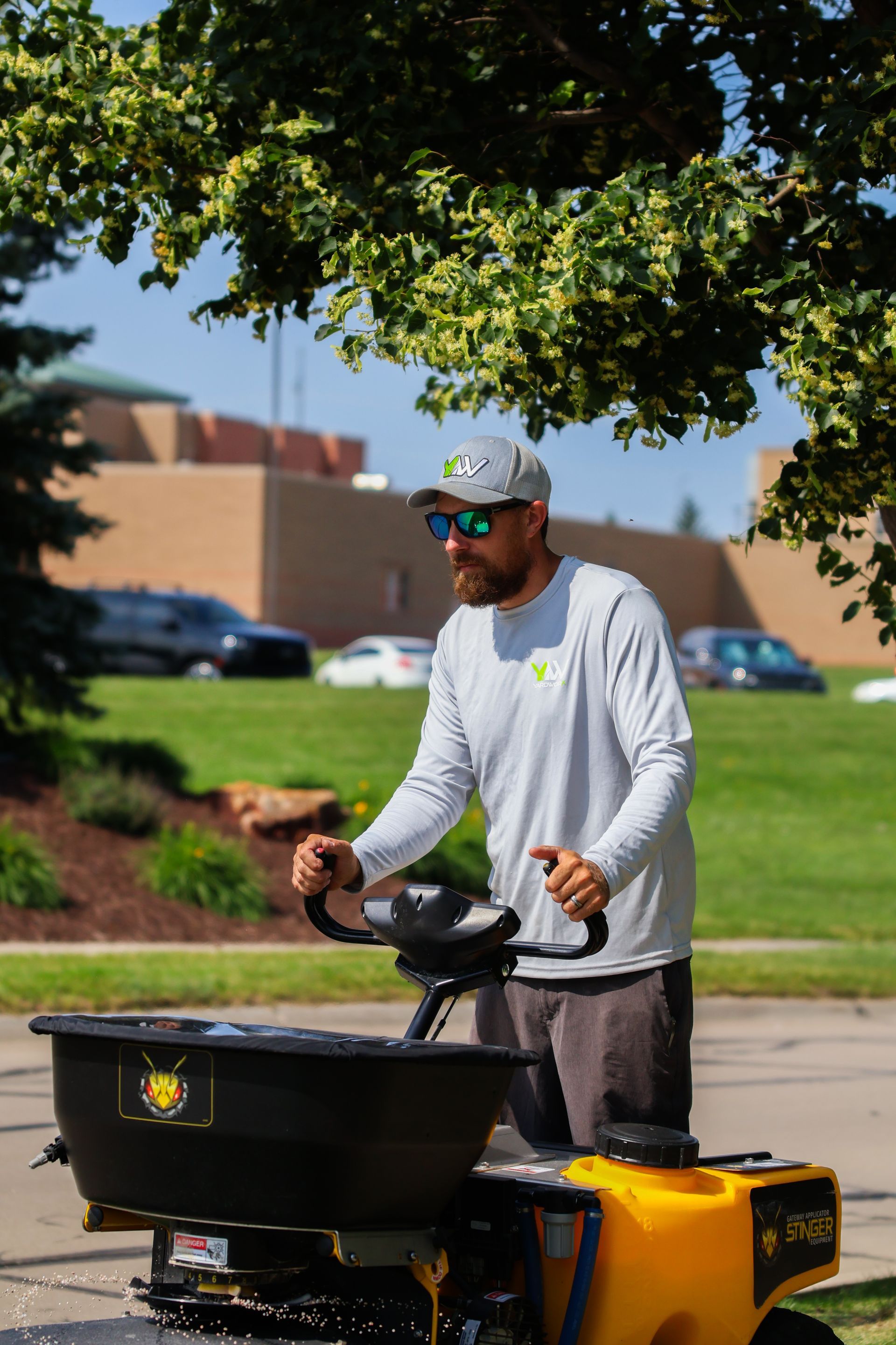 Man operating a yellow spreader, outdoors. He wears a hat, sunglasses, and a long-sleeved shirt. Sunny day.