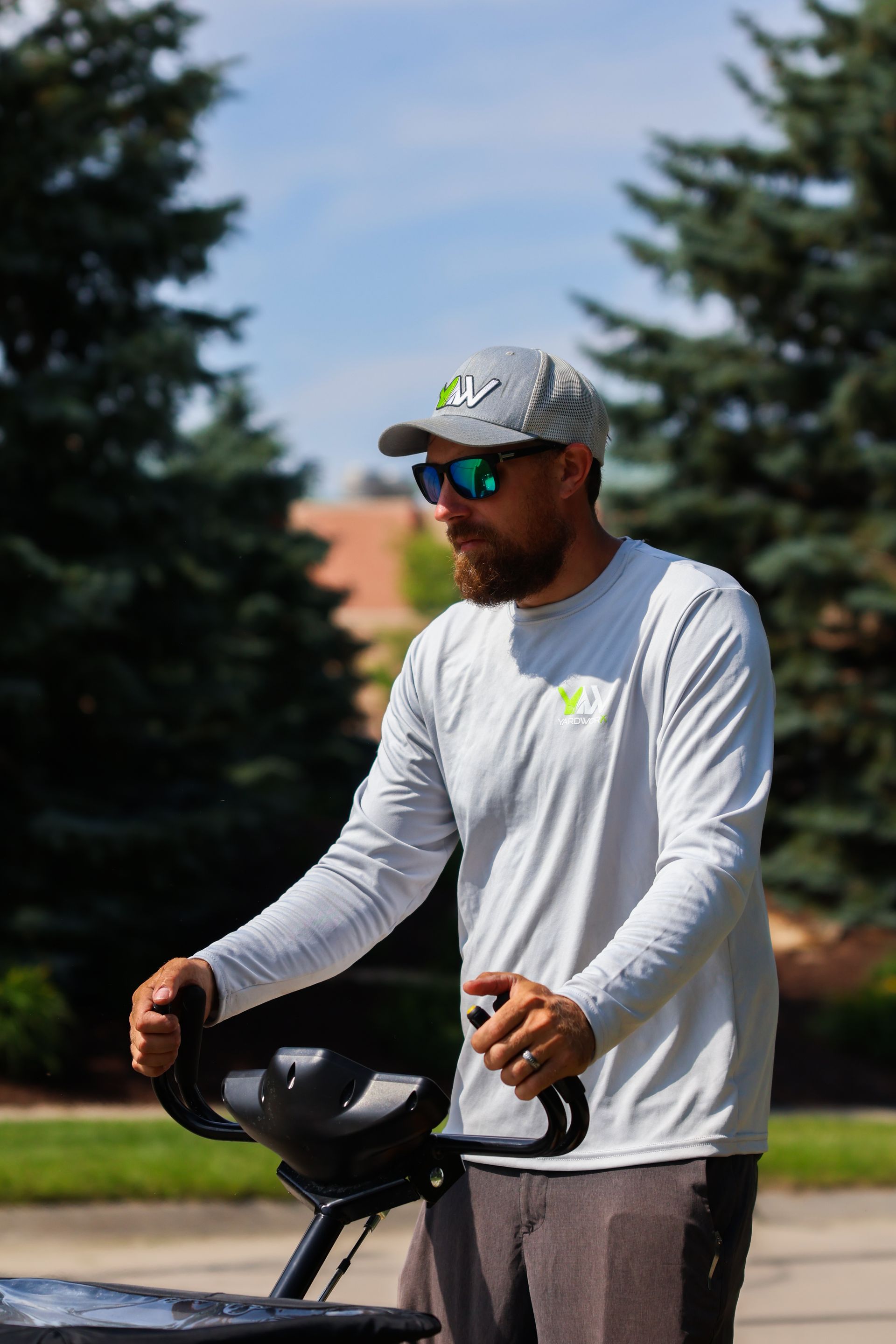 Man operating lawn care equipment outdoors, wearing sunglasses, hat, and long-sleeved shirt.