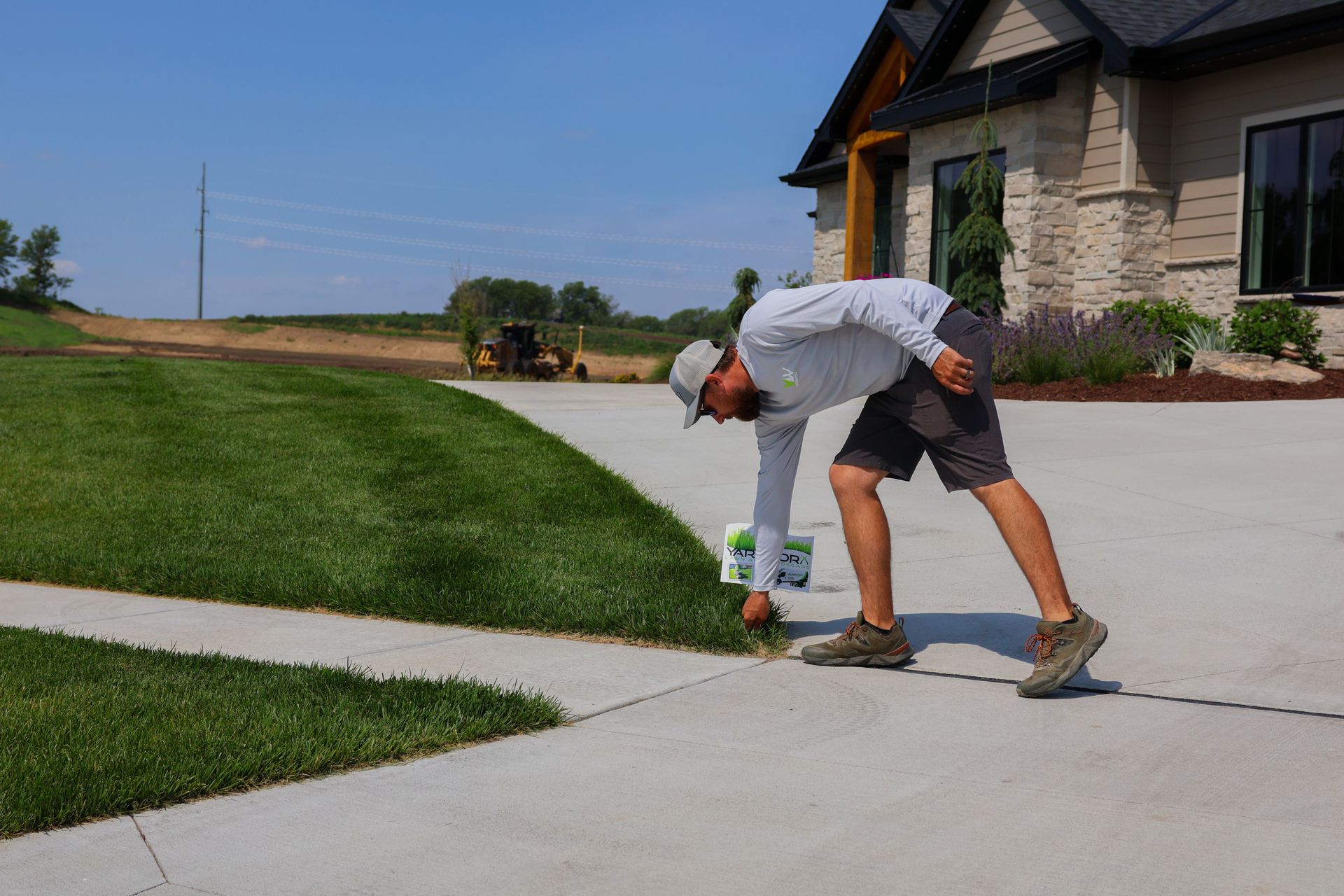 Man examining grass by a driveway, next to a manicured lawn and a house on a sunny day.