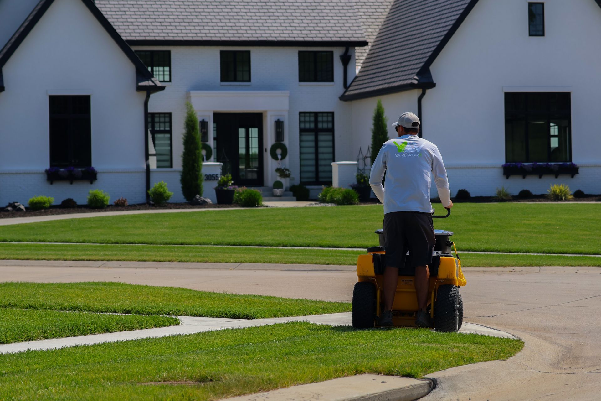 Person mowing a green lawn in front of a white house with black trim, using a riding mower.