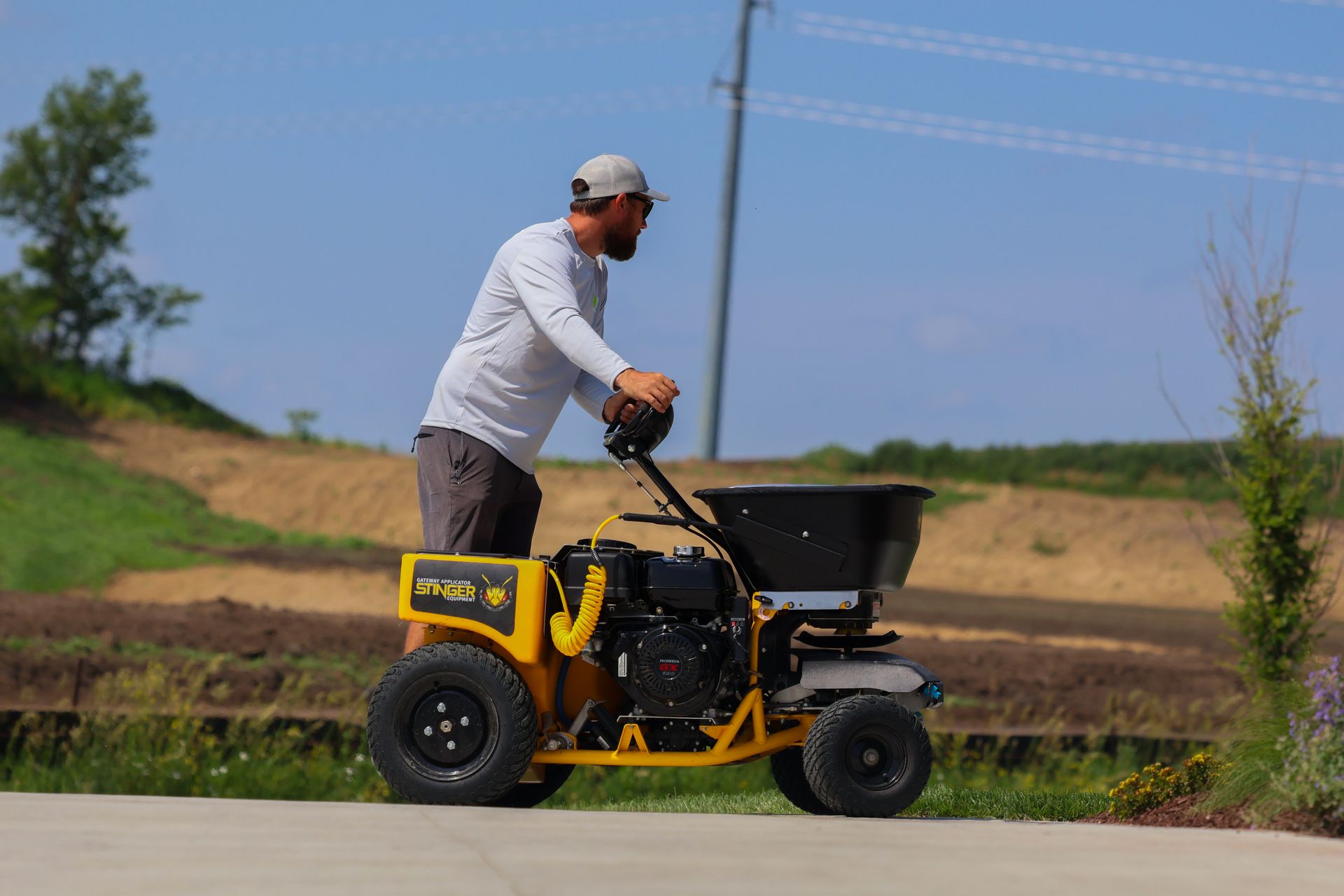 Man operating a yellow spreader on pavement, with a field in the background. Sunny day.