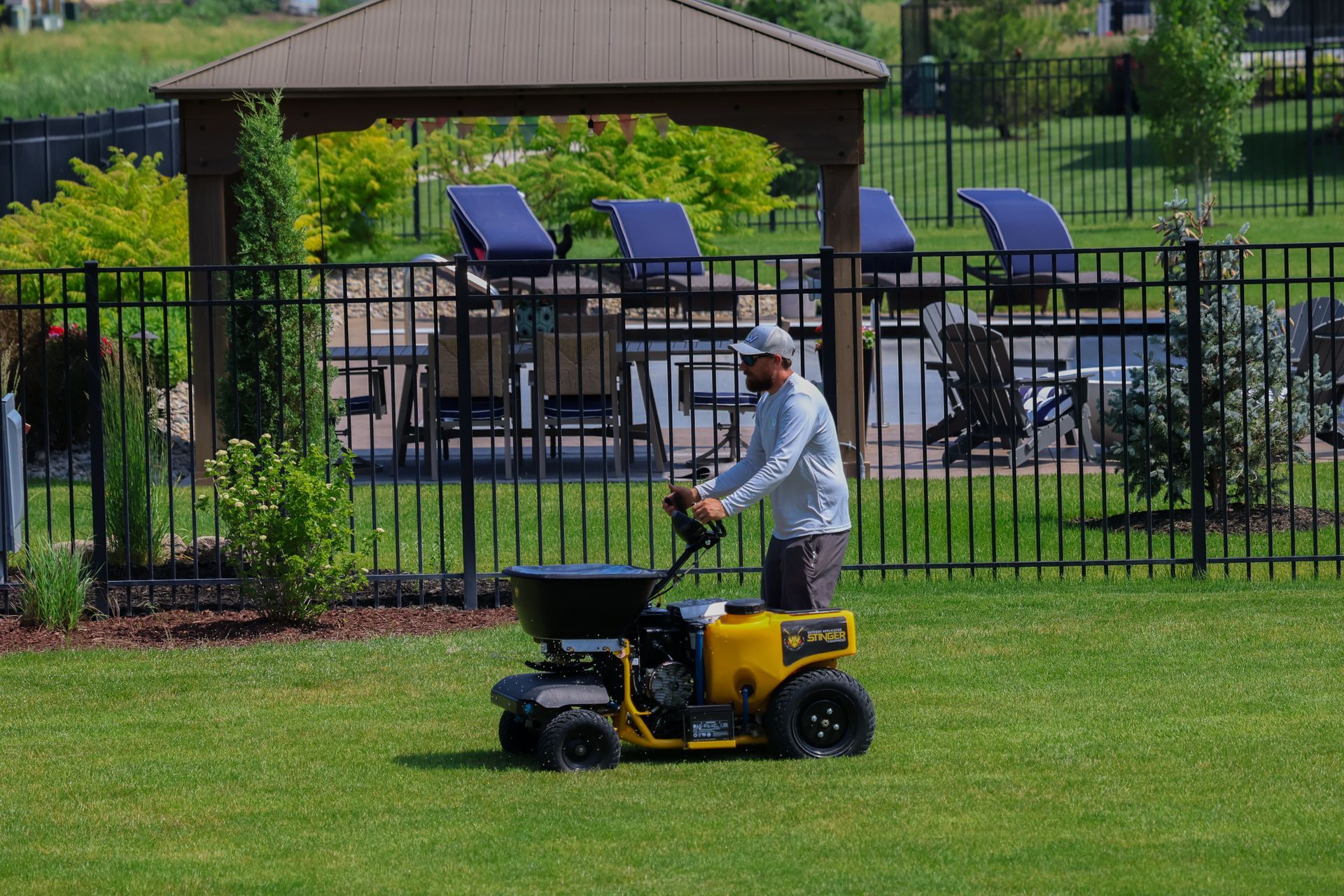 A person operating a yellow spreader on a lawn, near a black fence, gazebo, and pool.