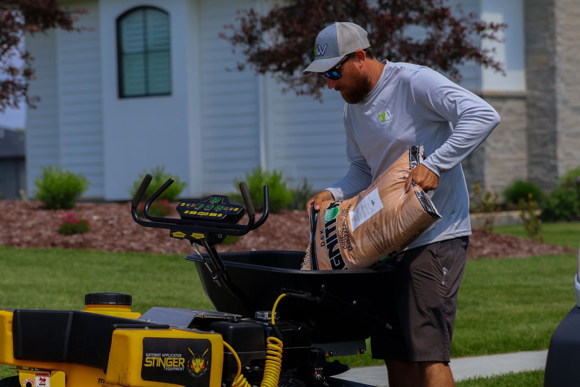 Man pouring contents from a bag into a lawn spreader. He wears a hat and sunglasses, standing in a yard.