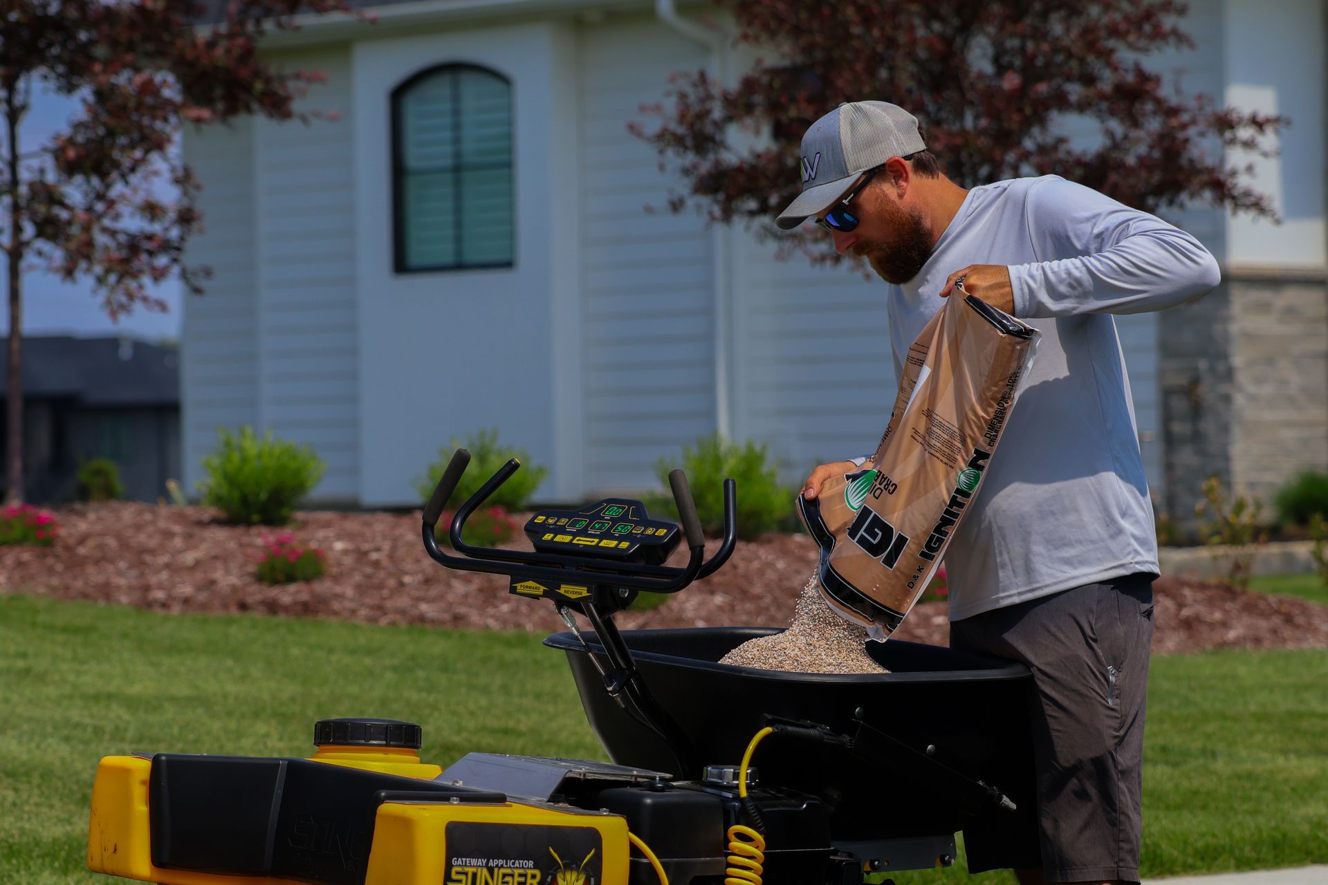 Man in sunglasses pouring seed into a spreader on a lawn. A house is in the background.