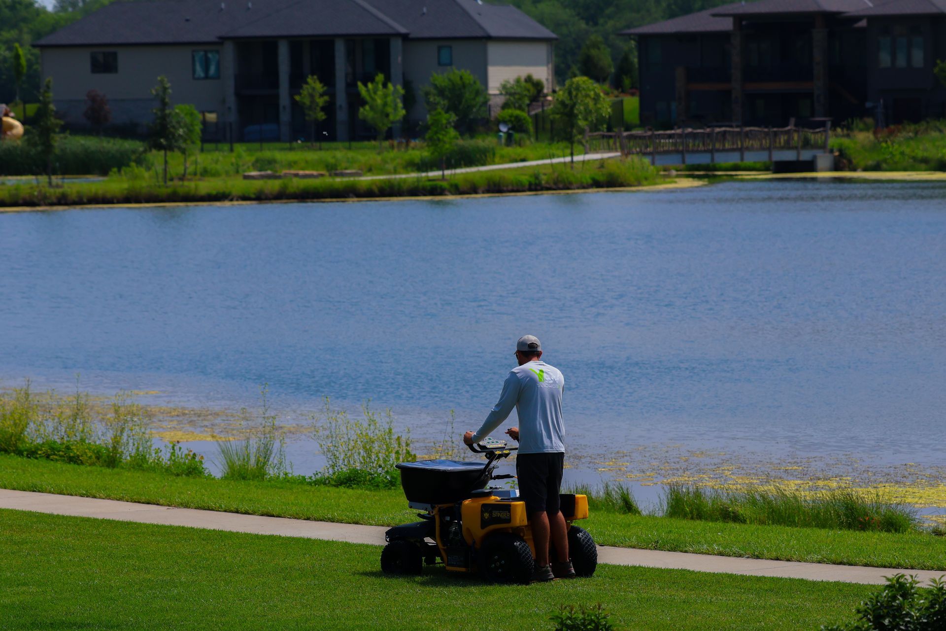 Man operating a yellow lawn spreader near a lake and houses.