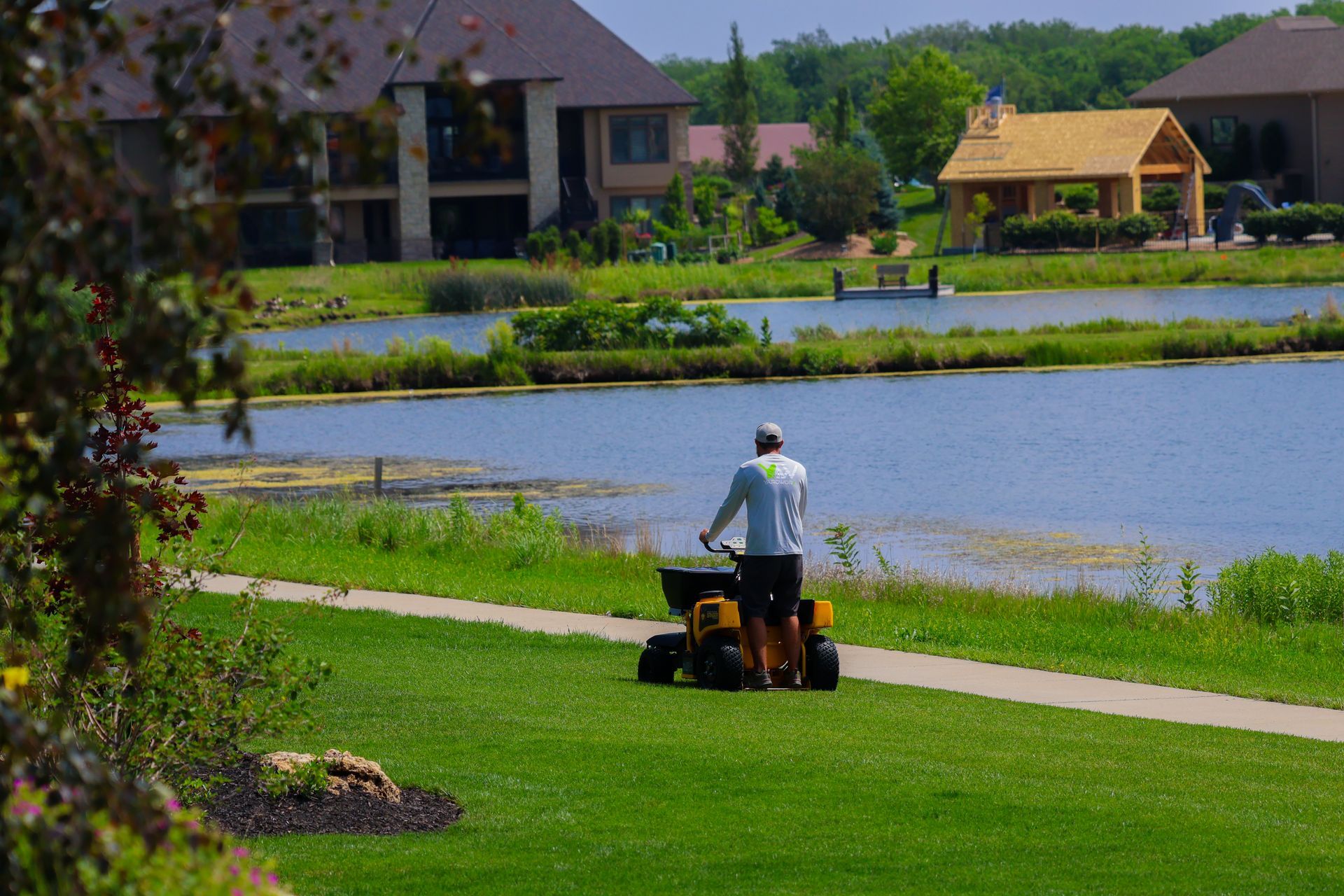 Man using a spreader on a grassy path near a lake, houses in background.