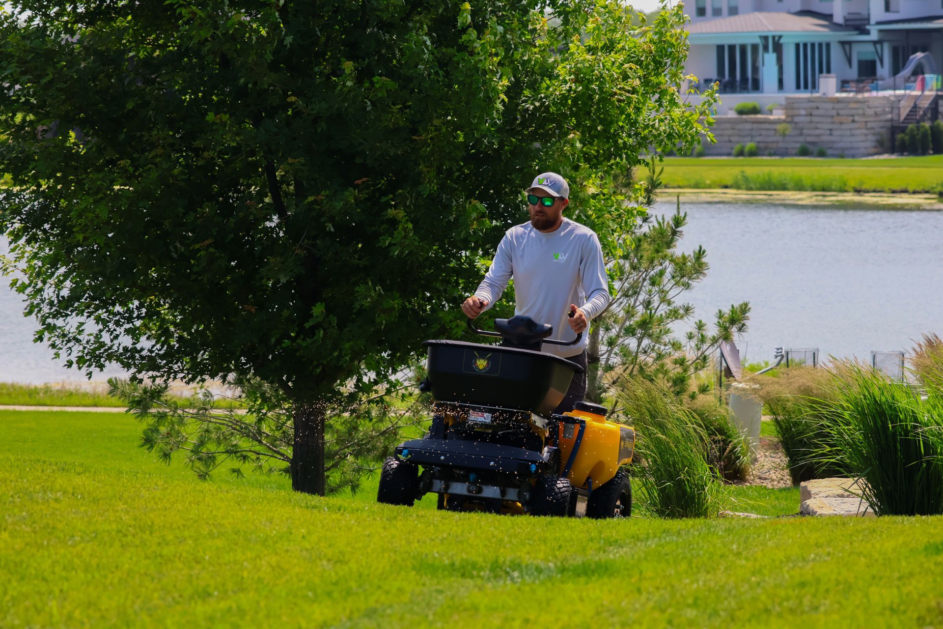 Man operating a yellow lawn spreader on a grassy hill near a lake.