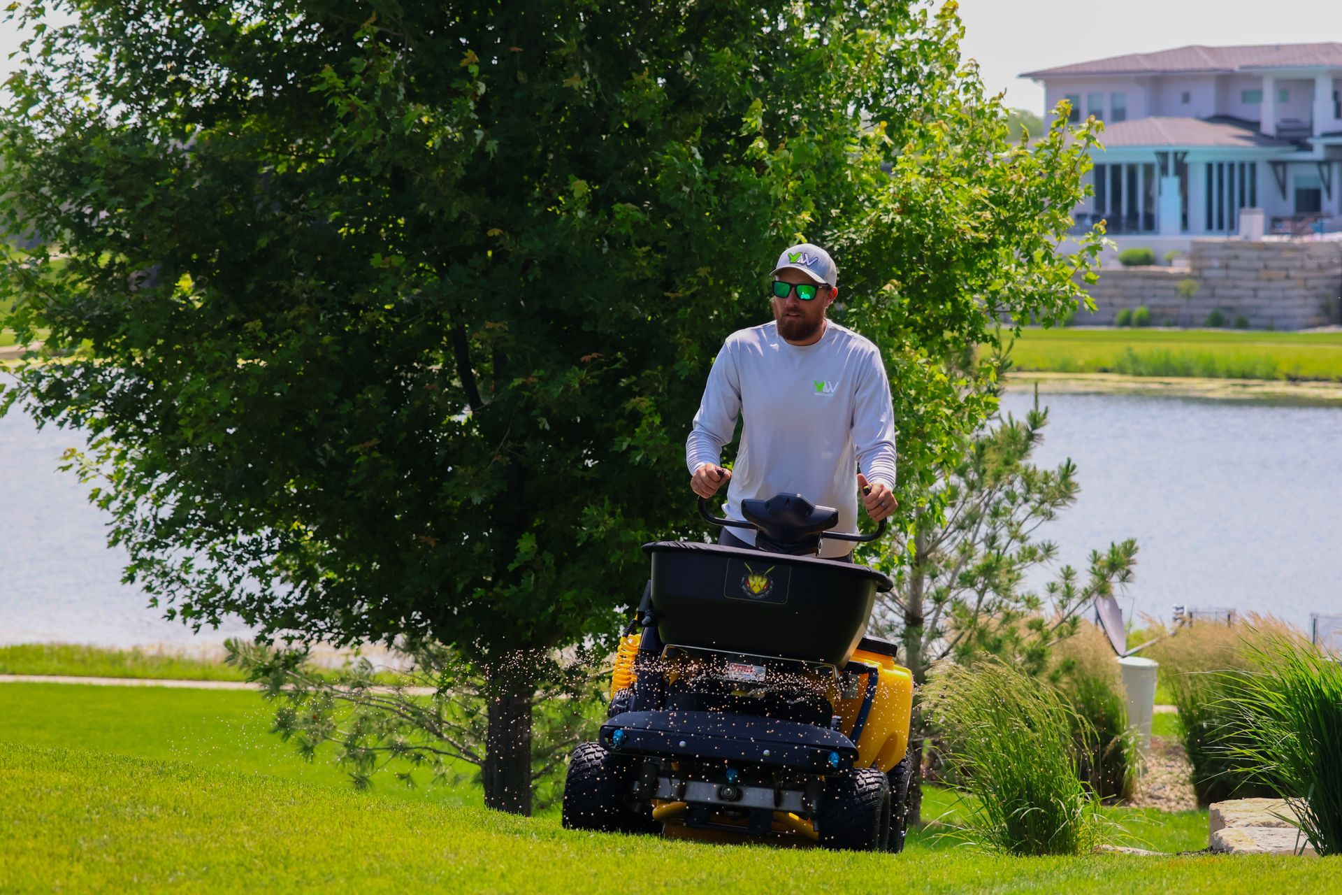 Man operating a yellow spreader on a grassy hill, near a lake.