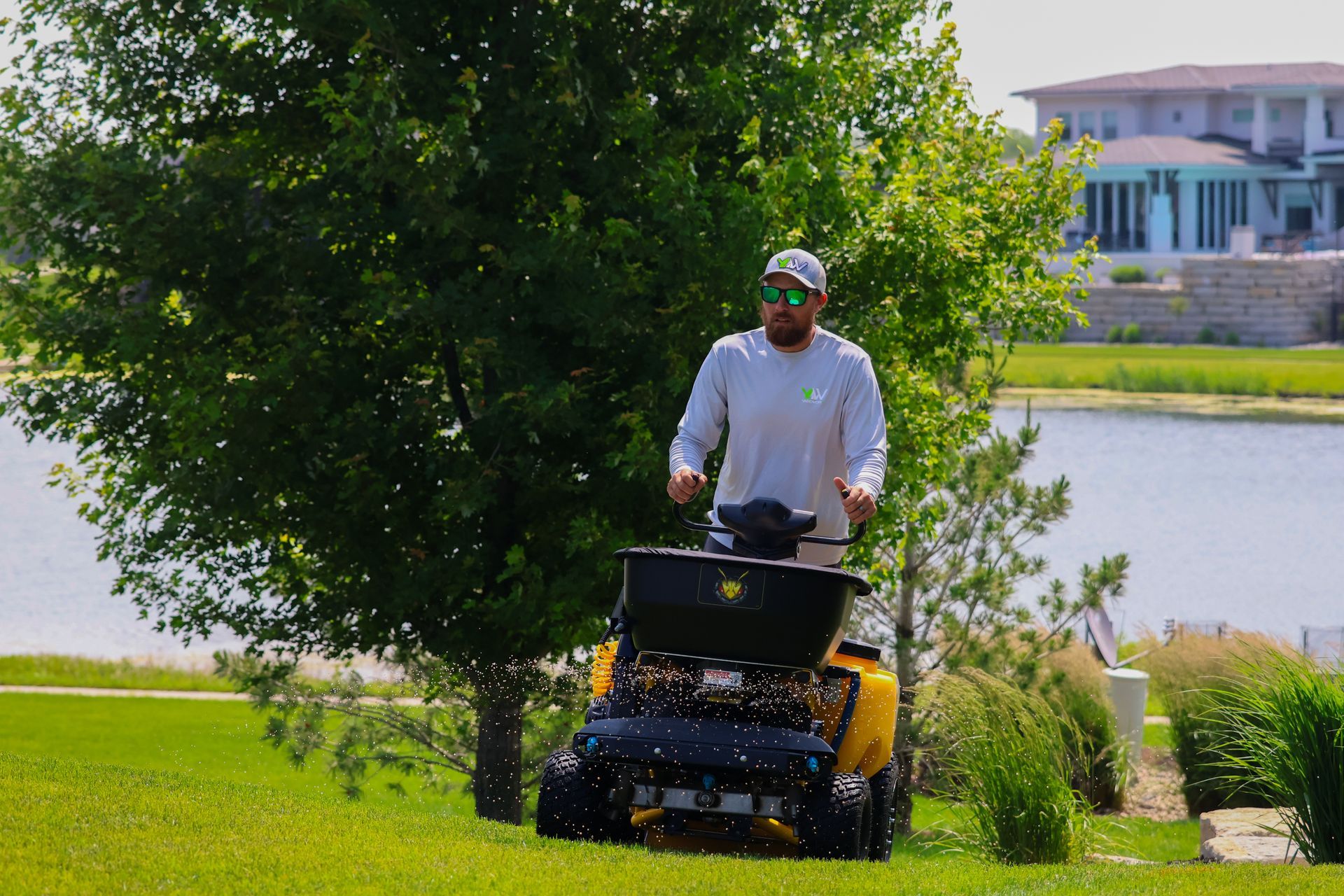 Man operating a yellow lawn spreader on a grassy hill near a lake.