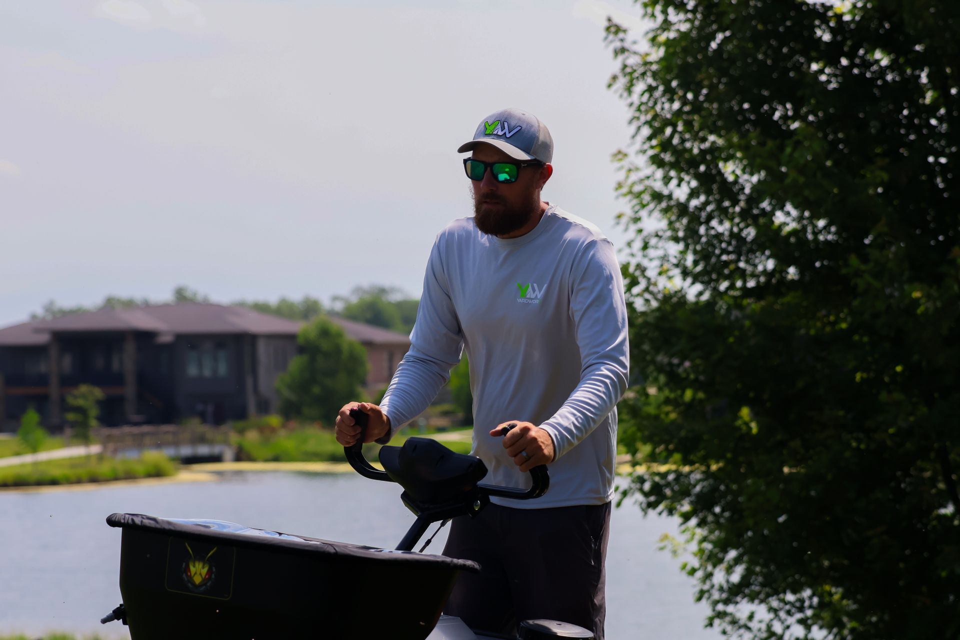 Man in sunglasses operating a spreader near a lake, wearing a hat and long-sleeved shirt.