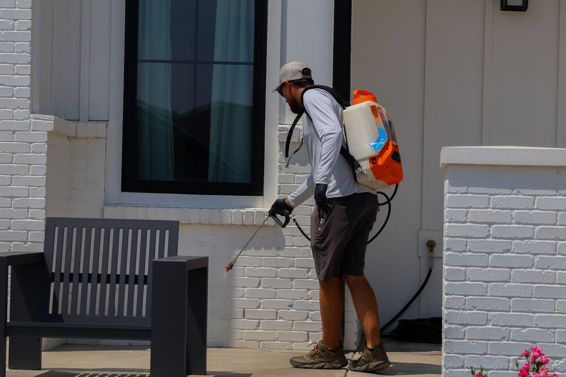 Man spraying insecticide near a white brick house, wearing a backpack sprayer, shorts, and hat.