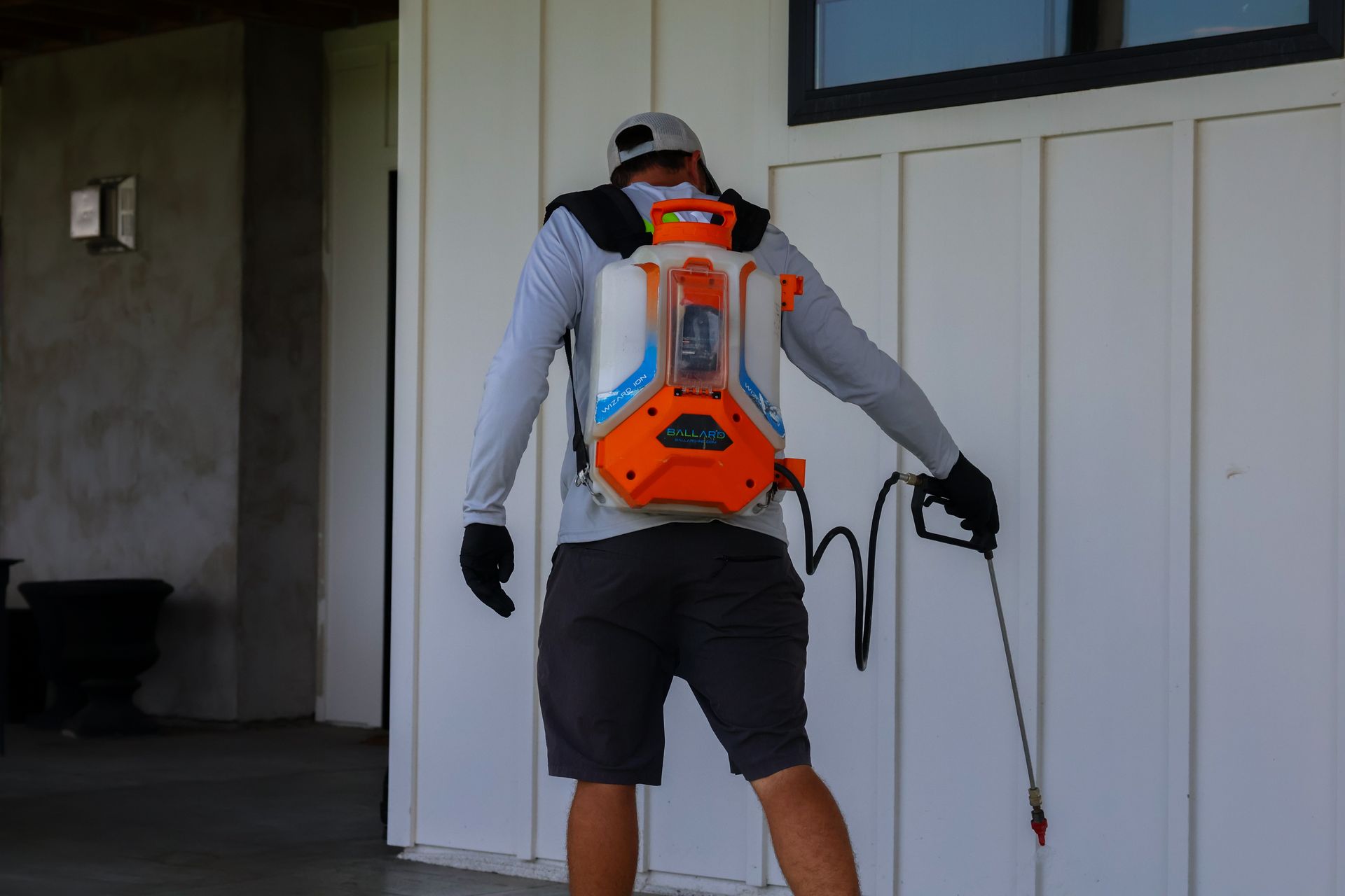 Person wearing sprayer backpack, spraying exterior wall. Orange, white, and black equipment.
