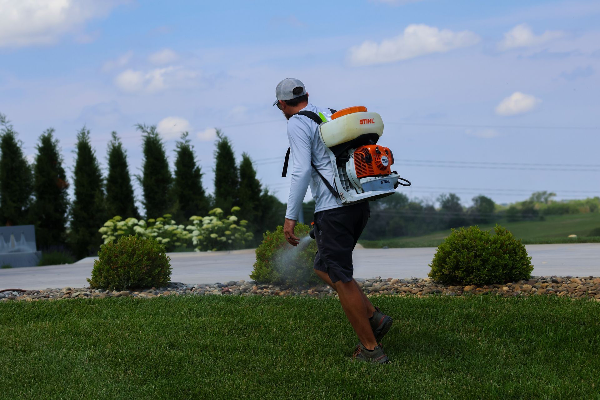 Person spraying lawn with backpack sprayer. Green grass, blue sky, and bushes in the background.