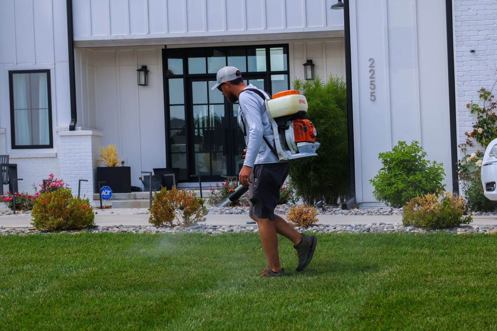 Man spraying lawn with backpack sprayer in front of a modern house.