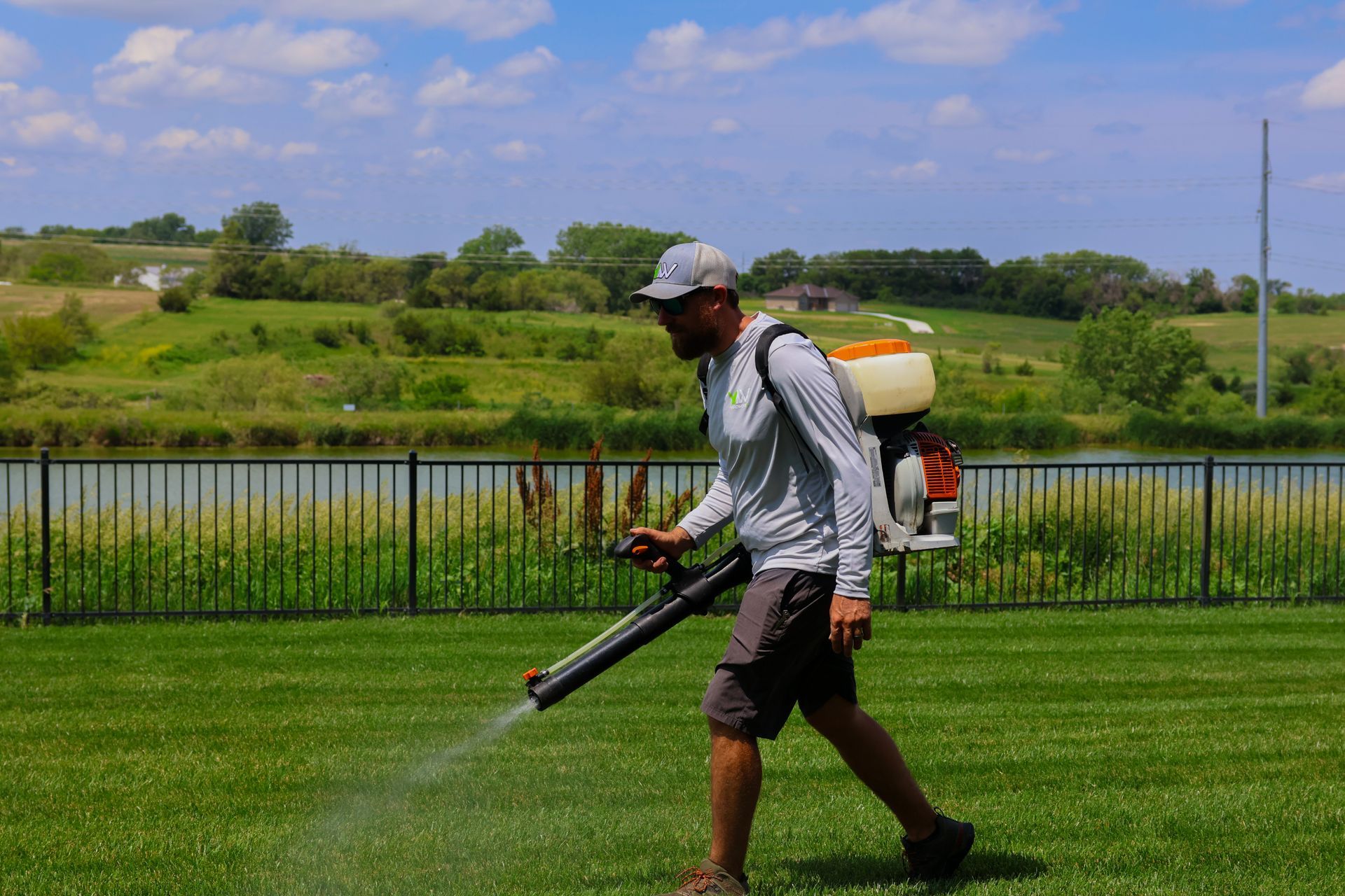 Man sprays lawn with a backpack sprayer in front of a lake and fence.