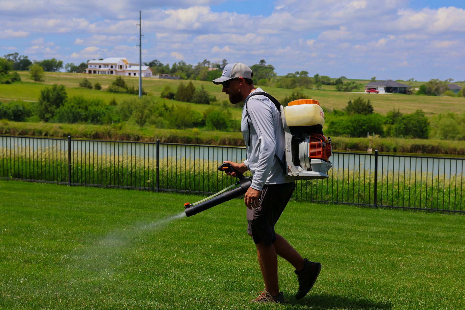 Man spraying lawn with backpack blower near a pond, sunny day.