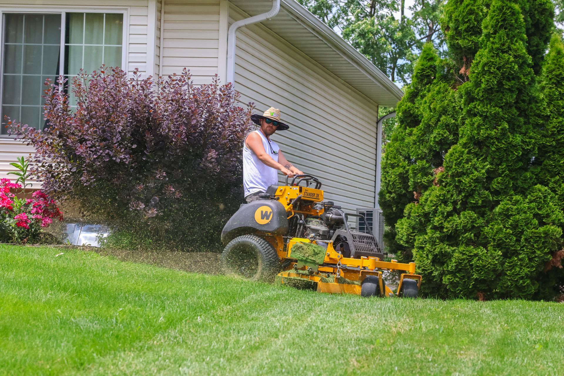 Person operating a yellow riding lawnmower, trimming grass next to a house with shrubs.
