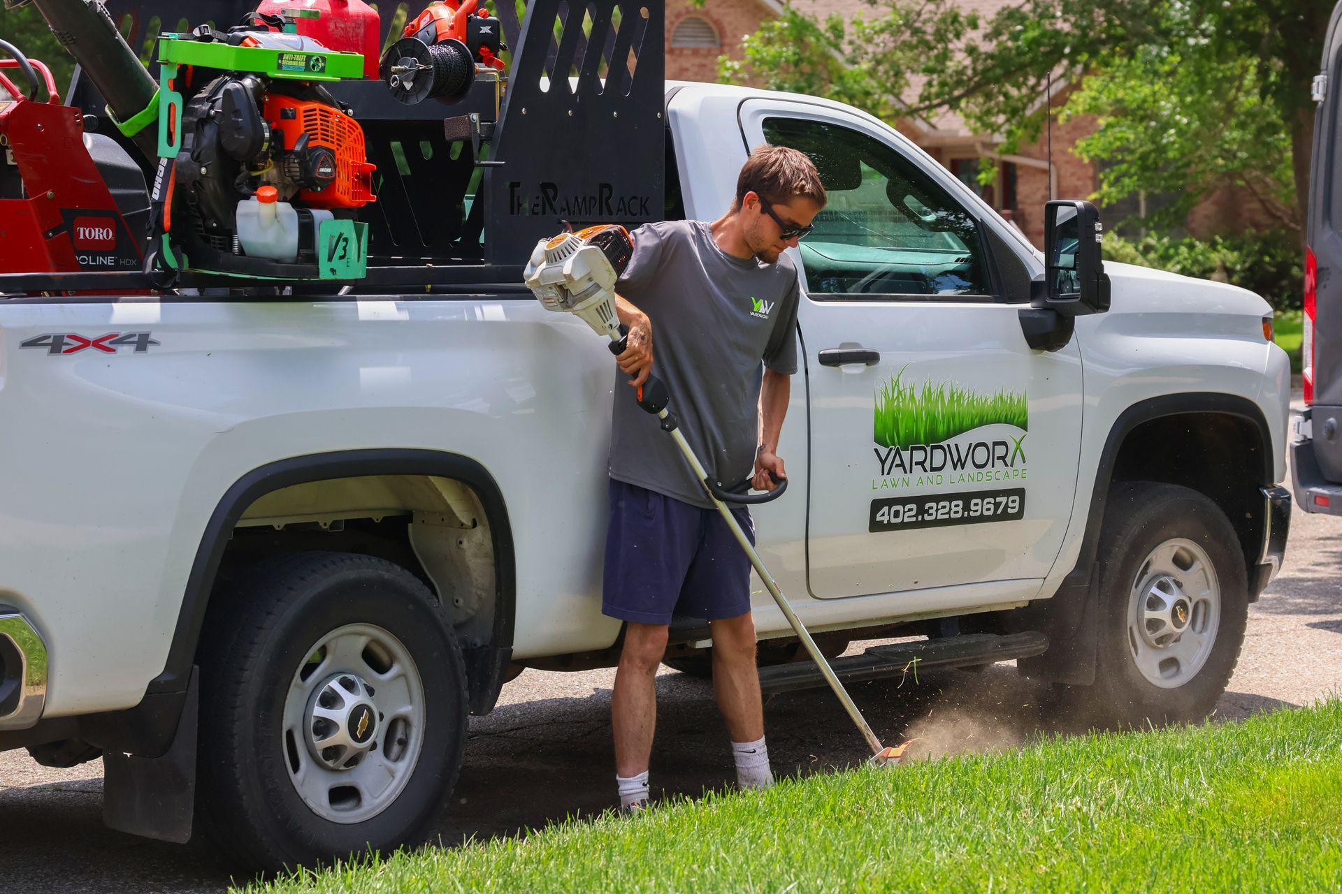 Man using a string trimmer to cut grass near a white truck with landscaping equipment.